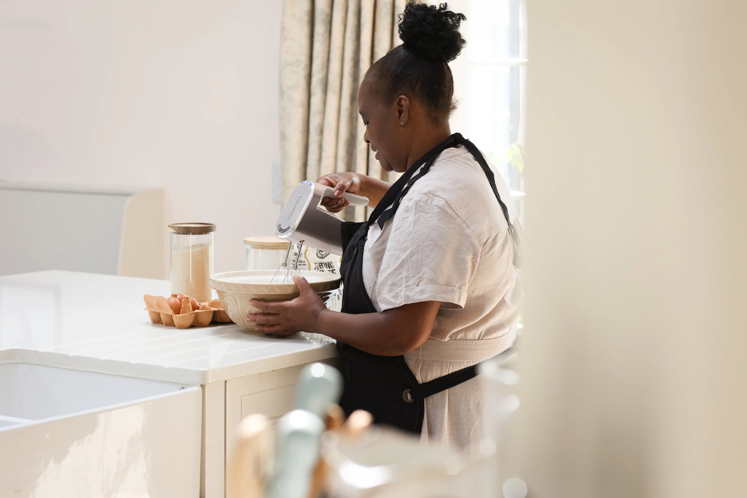 Woman in casual clothing and apron mixing ingredients with a hand mixer in a kitchen.