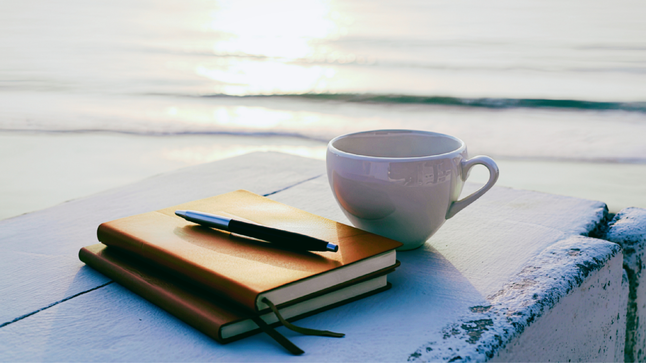 A white coffee mug, a stack of two notebooks with a pen on top, and a piece of coral on a white surface outdoors by the water during sunset.