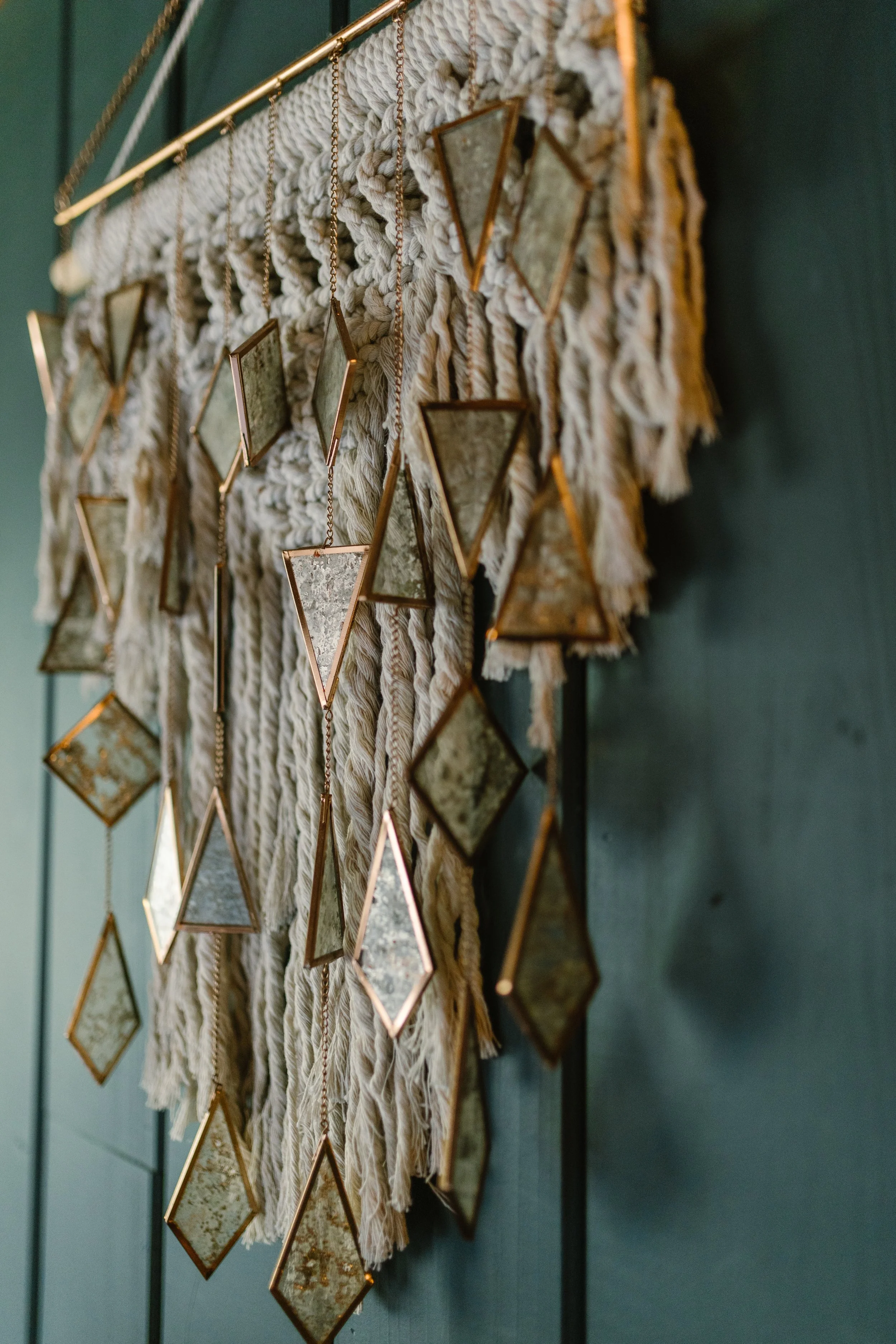 Wall hanging art piece consisting of a frayed beige fabric background with framed mirrors and geometric shapes hanging from chains.