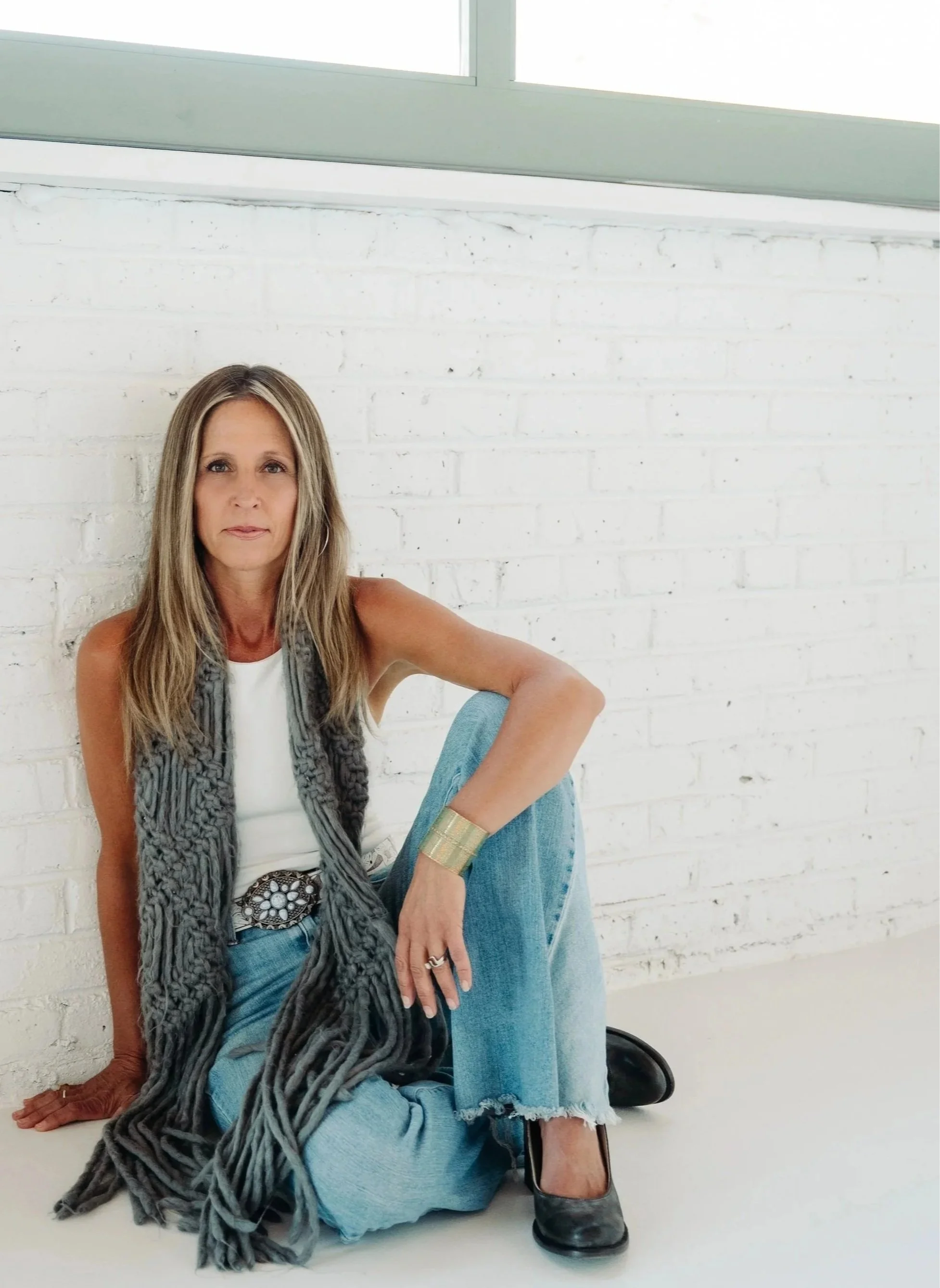 A woman with long blonde hair, sitting on the floor against a white brick wall, wearing a grey scarf, a white top, ripped blue jeans, and black boots, with jewelry on her left hand and wrist.