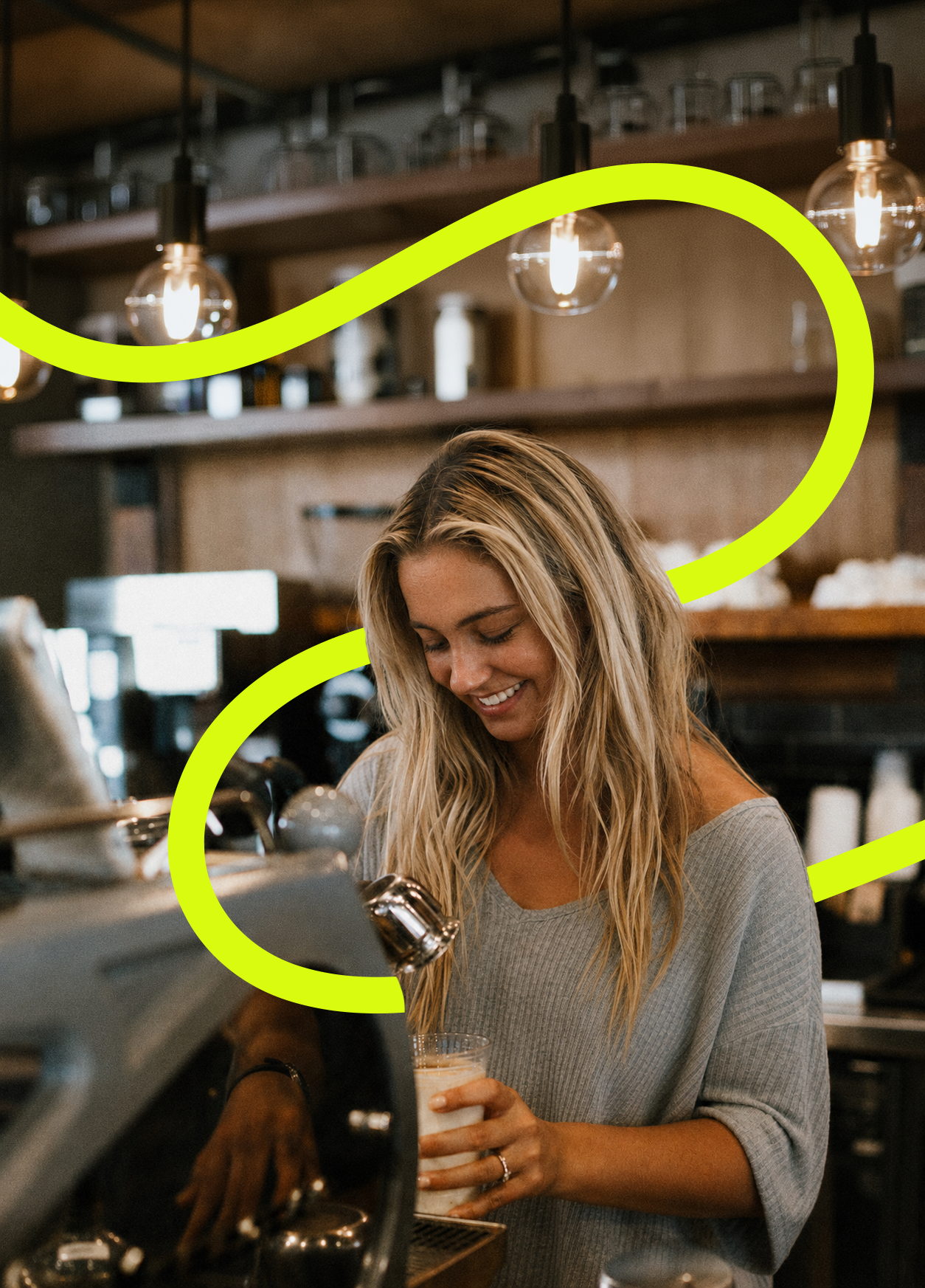 A young woman with long blonde hair smiling as she holds a glass of coffee or latte at a coffee shop counter.