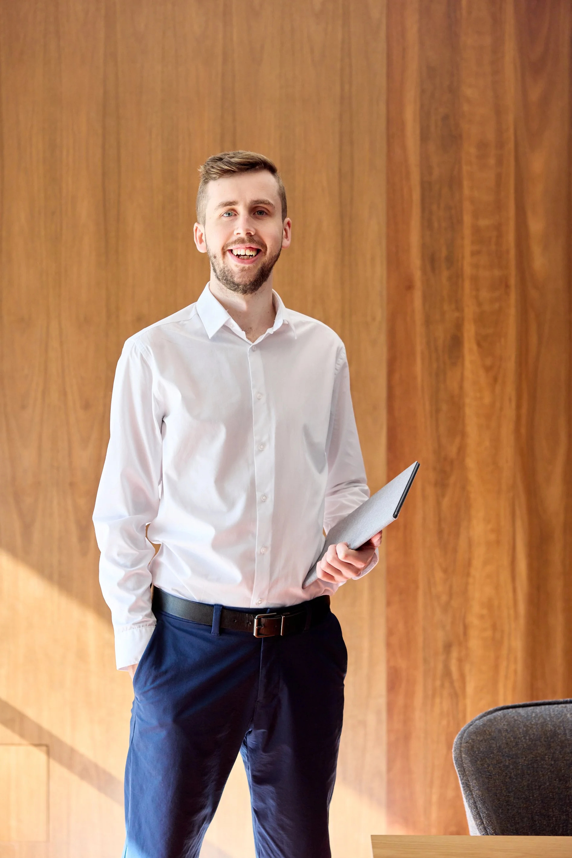 A young man with light skin, short brown hair, and a beard, wearing a white dress shirt and dark trousers, smiling and holding a folder, standing in front of a wooden wall in a professional setting.