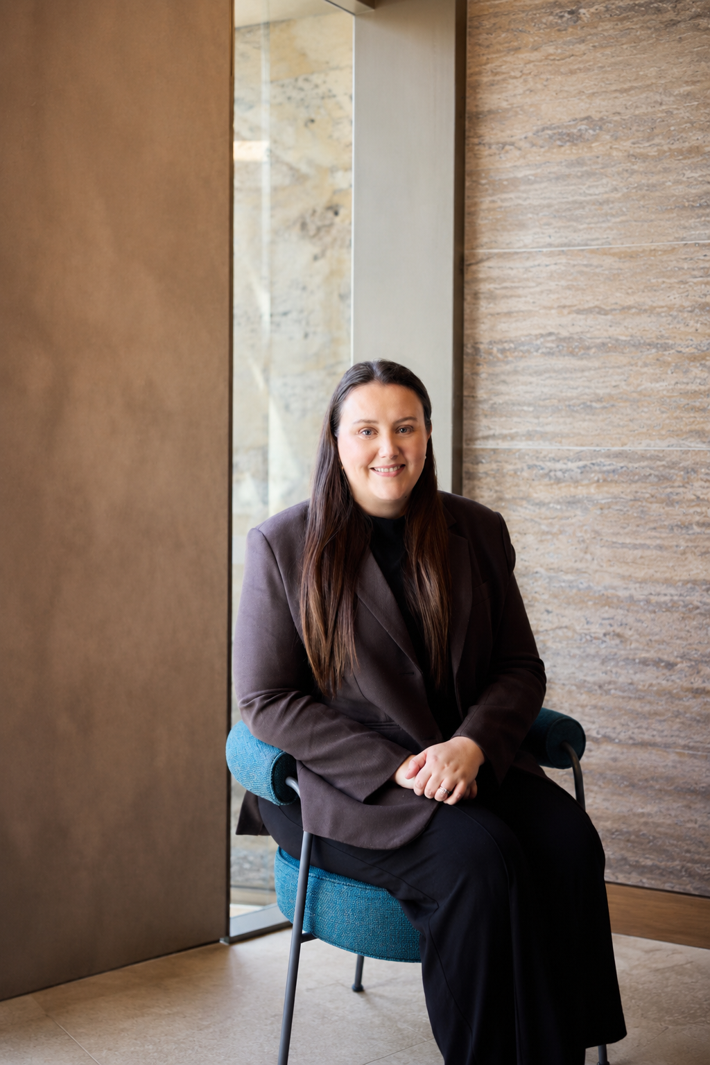 A woman with long dark hair, wearing a dark blazer and black pants, sitting on a blue chair in a modern indoor setting with textured stone walls and a large window in the background.
