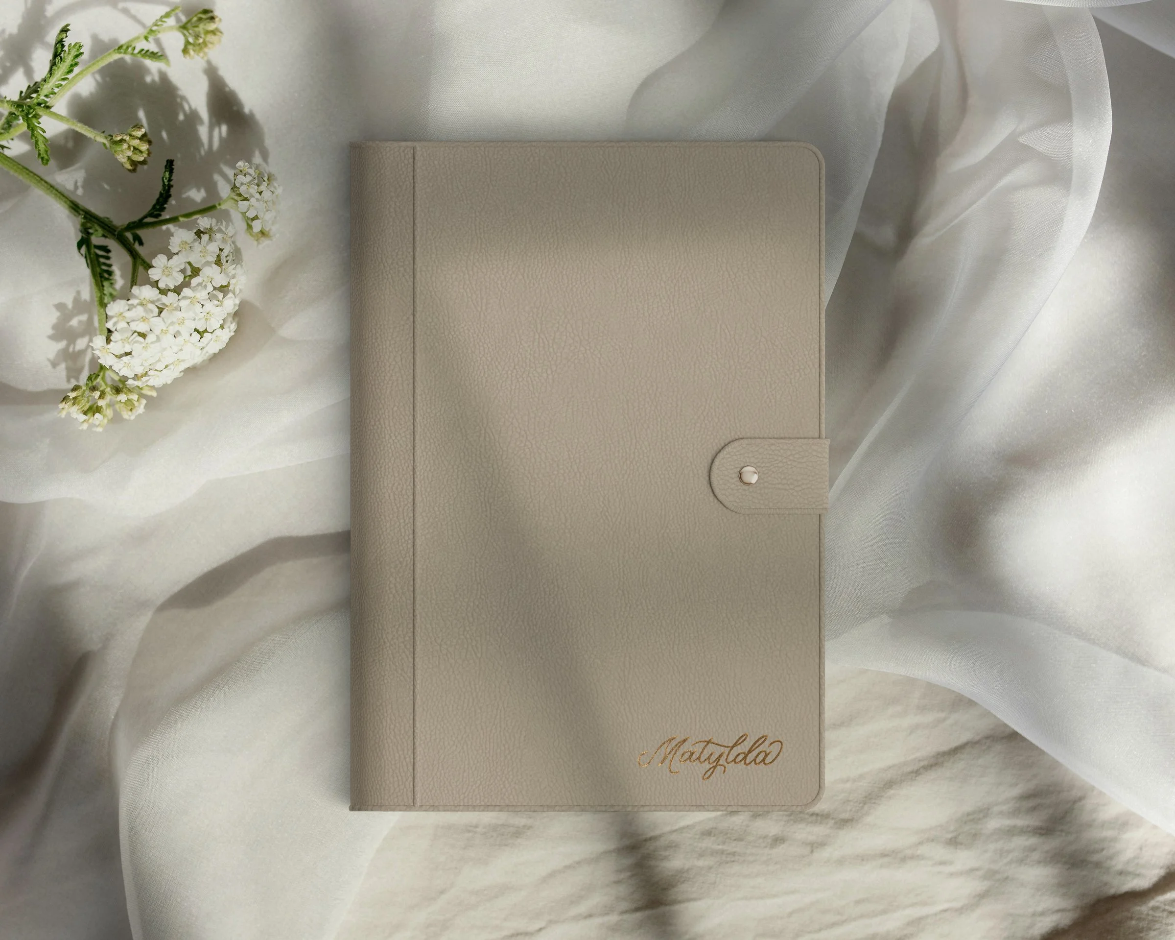 Soft taupe leather-bound journal with snap closure, embossed with Matylda in gold foil script in the bottom right corner. It rests on beige and white flowing fabric with white flowers to the left, lit by sunlight with soft shadows for depth.