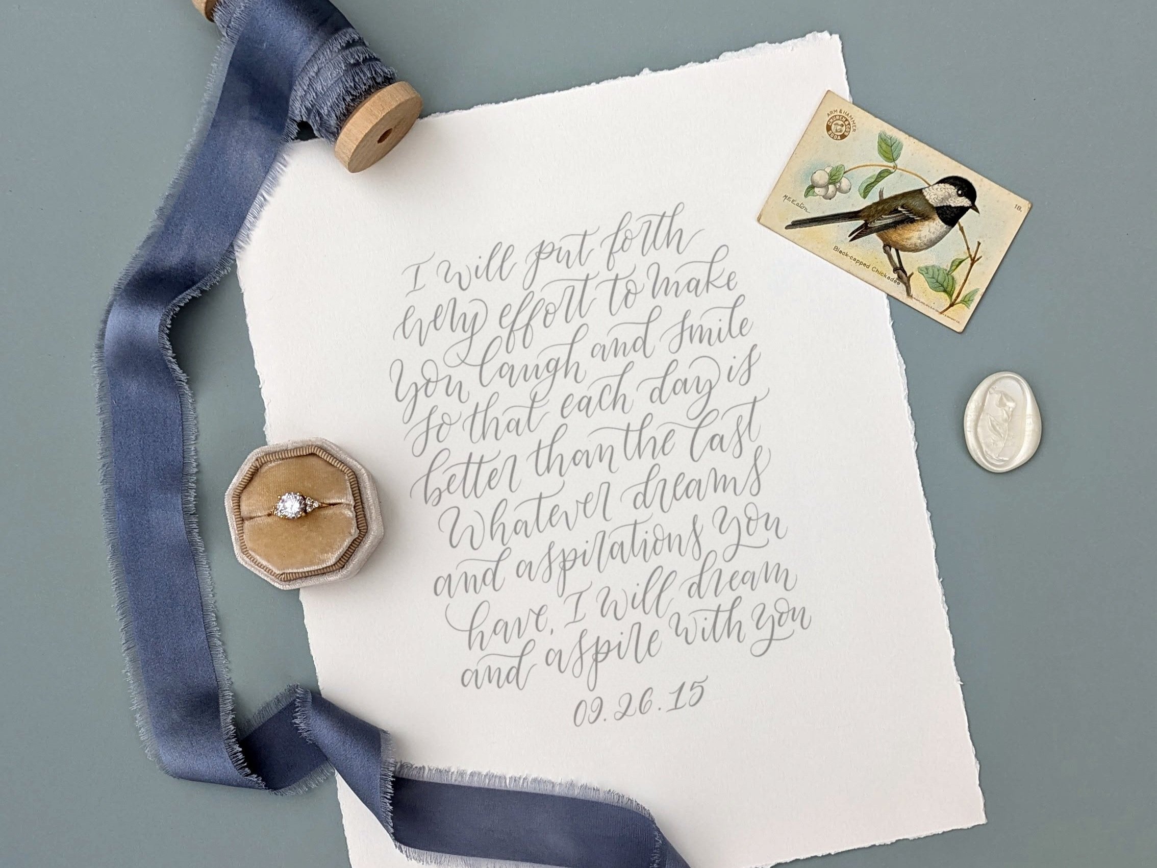 Flat lay of a handwritten love note in elegant gray calligraphy on torn-edge paper, reading a heartfelt vow dated 09.26.15. Styled with dusty blue ribbon, velvet ring box holding a diamond ring, a bird postcard, and a wax seal on a muted gray surface