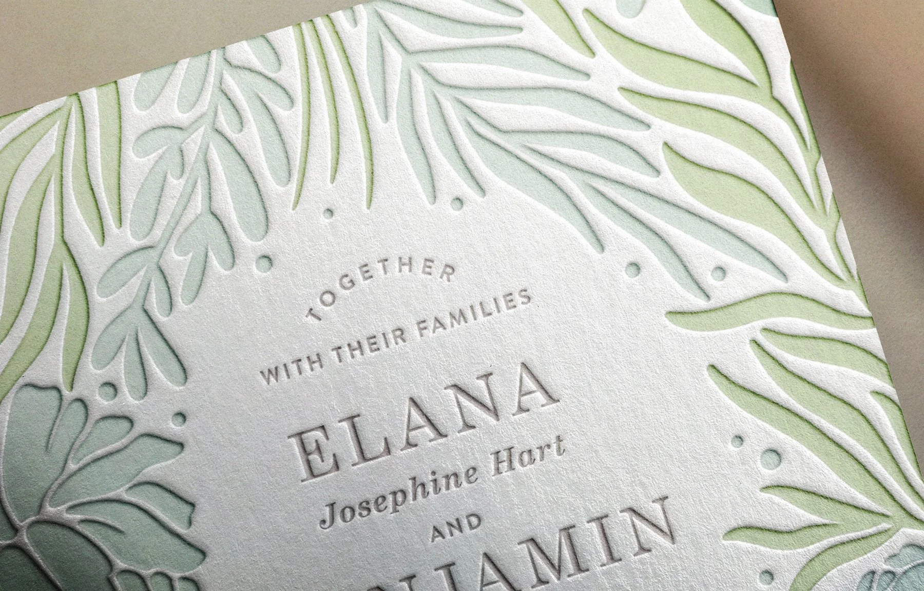Closeup of a textured wedding invitation with leafy cutout details in soft green and blue  featuring names Elana Josephine Hart and Benjamin in soft gray with debossed lettering and refined typography Merges elegance with natural design elements