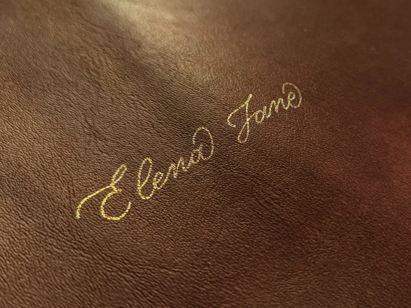 Close-up of textured brown leather with the name Elena Jane elegantly embossed in shimmering gold cursive script capturing the luxurious detail and custom personalization of the surface. Shows fine creases and soft lighting creating warm rich feel.