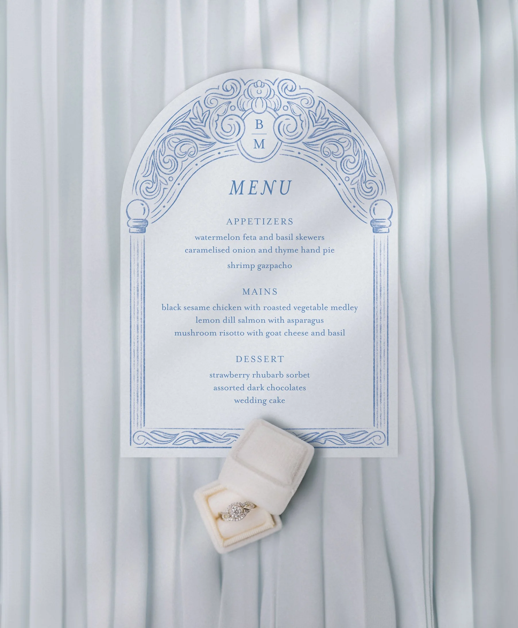 Elegant wedding menu card with blue ornamental arch on pillars and initials BM at the top featuring three courses in centered alignment. Card rests on crisp white pleats, with a velvet ring box containing a diamond engagement ring below it.