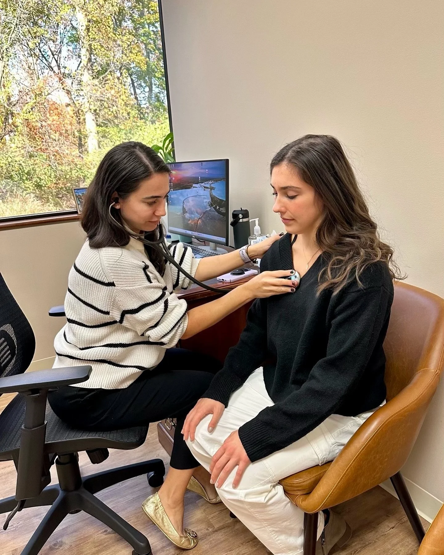 A female healthcare provider uses a stethoscope to listen to a woman's chest in a medical office with a window showing trees outside.