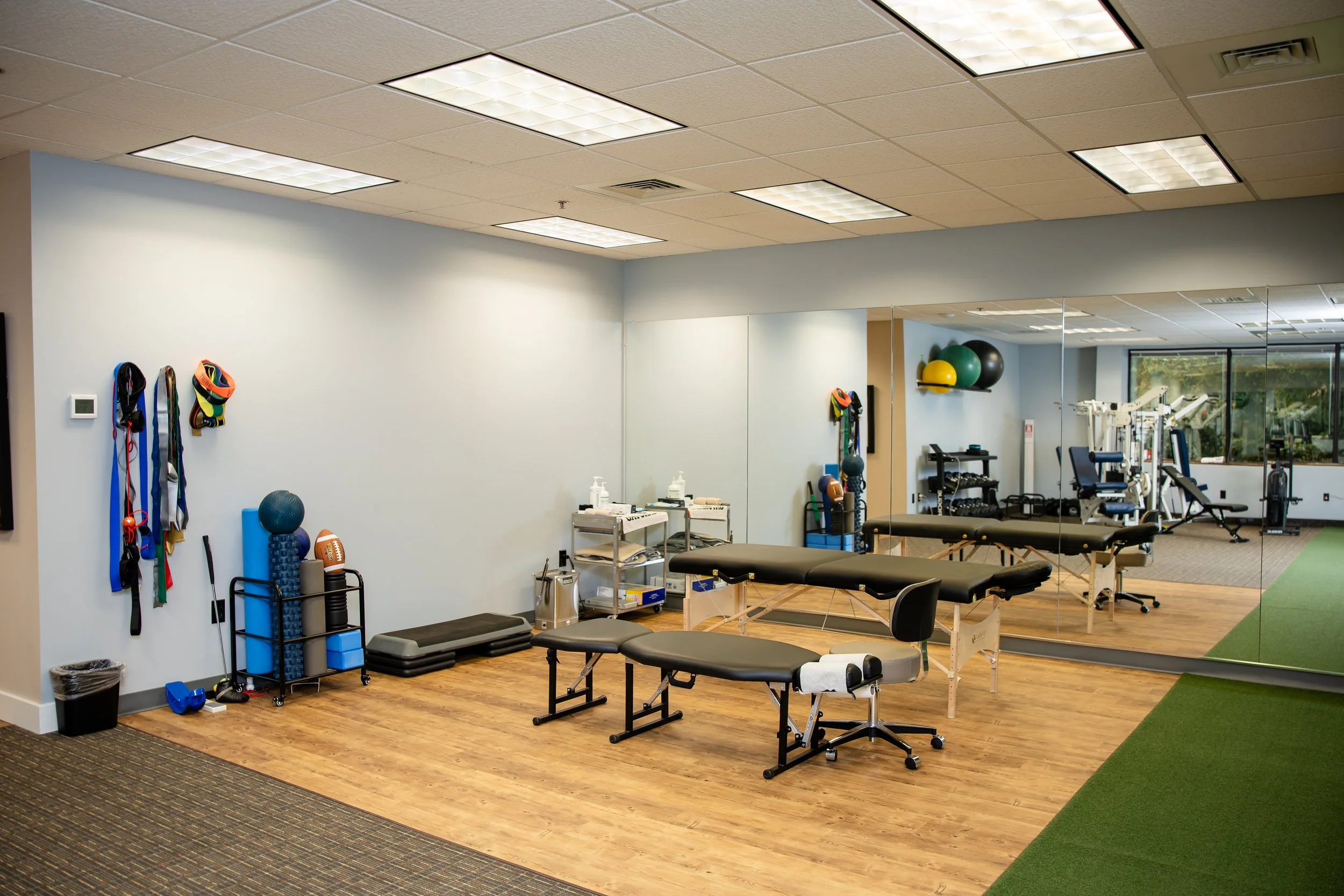 A physical therapy or fitness room with treatment tables, exercise balls, resistance bands, dumbbells, and mirrors.