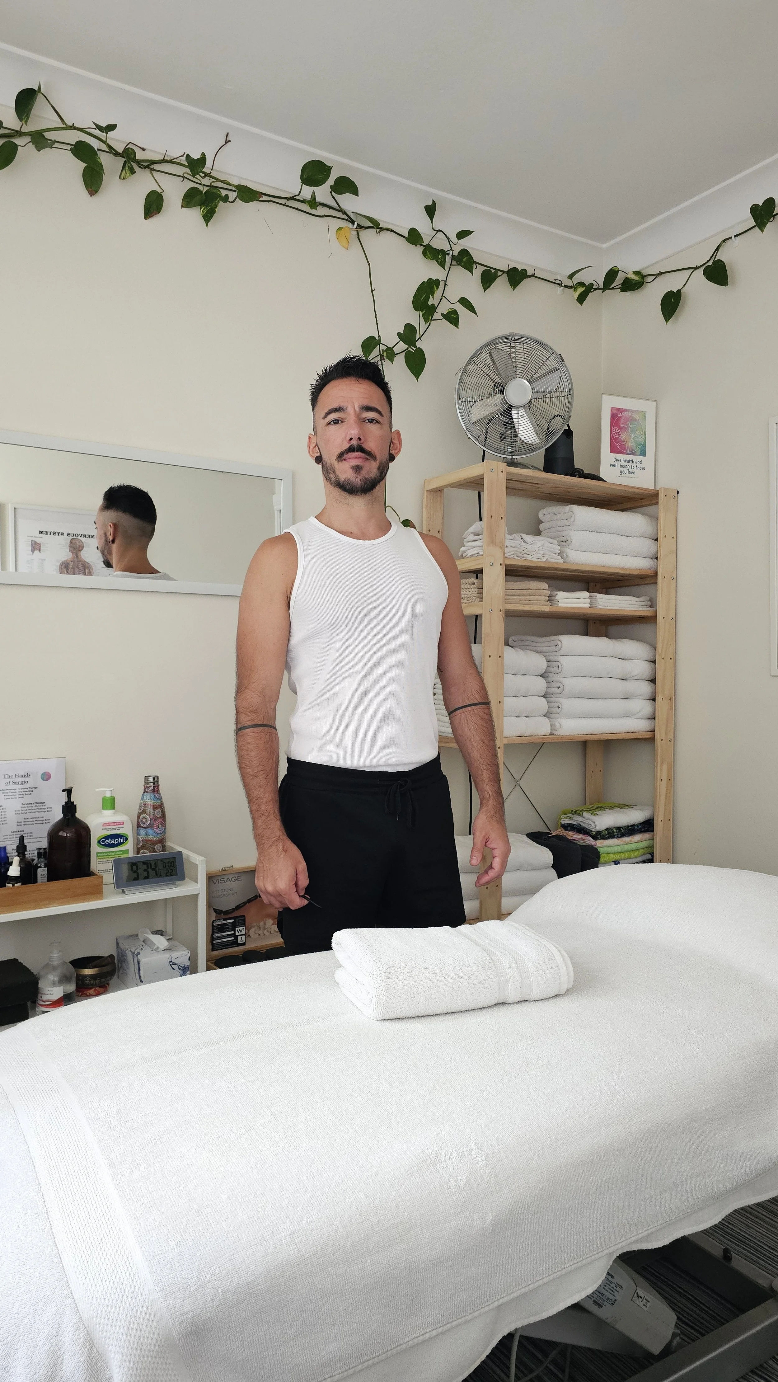 A man standing in a wellness or massage room, facing the camera, with a massage table in the foreground, towels on shelves behind him, and a mirror on the wall.