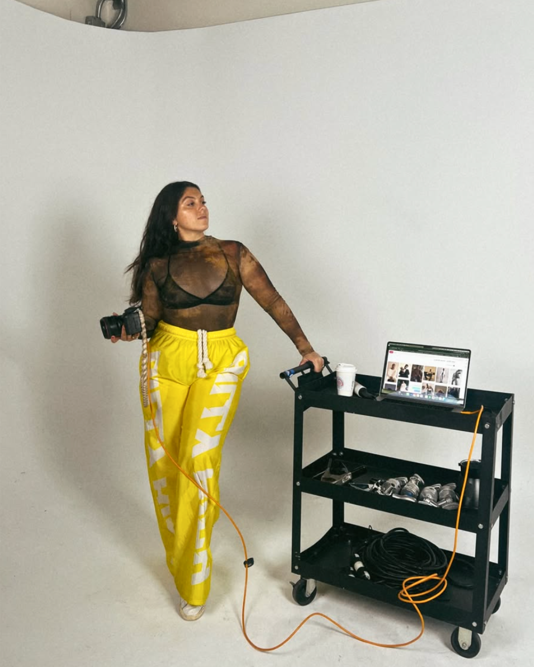 Woman in black mesh top and yellow sweatpants with white letters, holding a camera and leaning on a black cart with a laptop, coffee cup, and various filming equipment