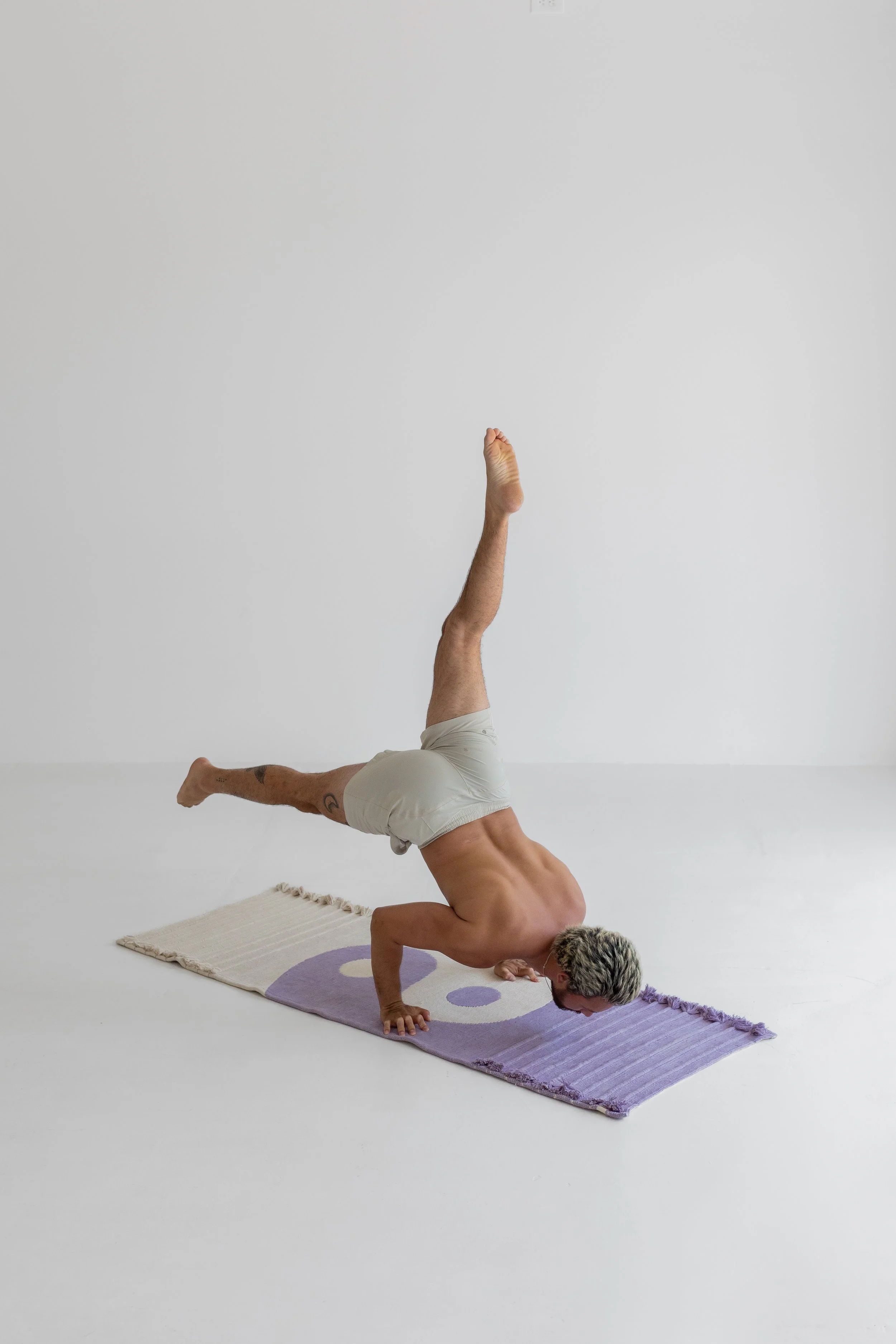 A shirtless man with gray hair practicing yoga in a headstand position on a purple and cream rug in a plain white room.