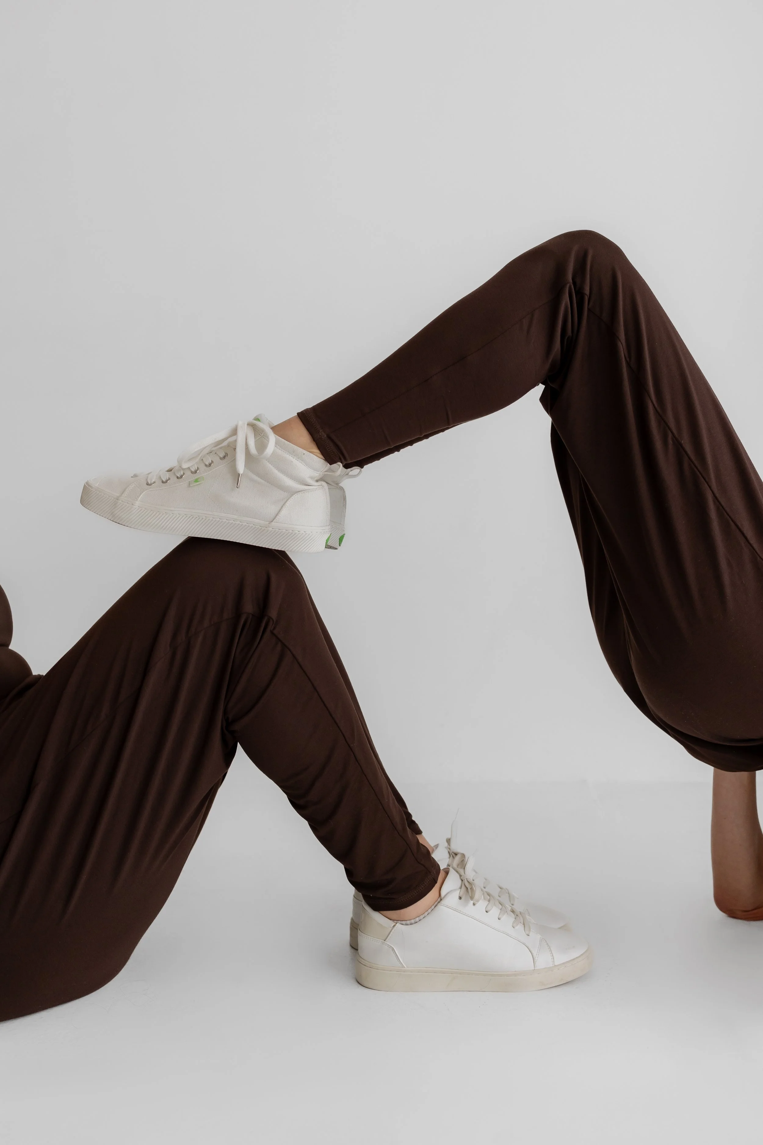 Person lying on the ground with legs raised, wearing brown pants and white sneakers, against a plain white background.