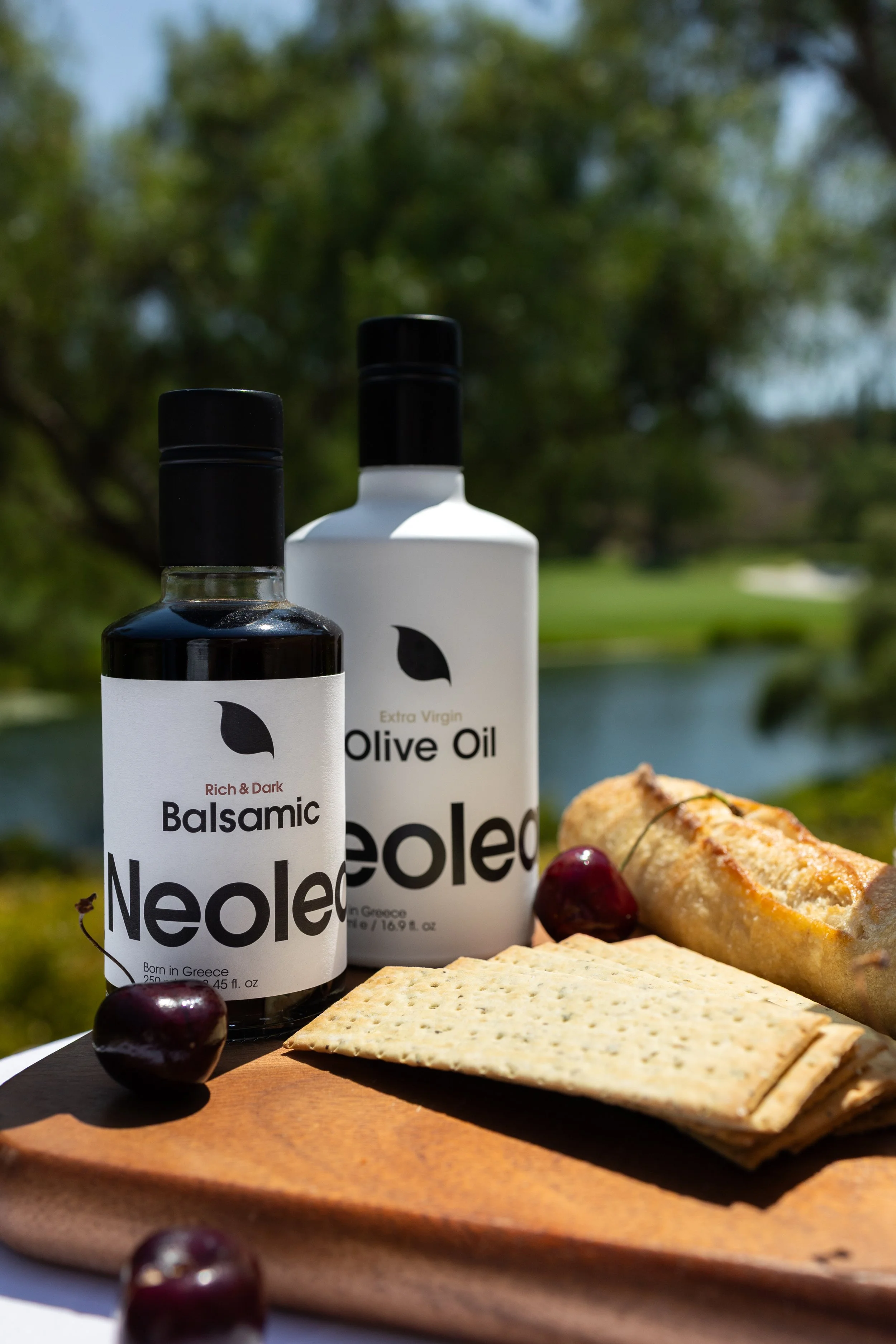 Bottles of balsamic vinegar and olive oil on a wooden cutting board with crackers, cherries, and a pastry in an outdoor setting with a lake and trees in the background.