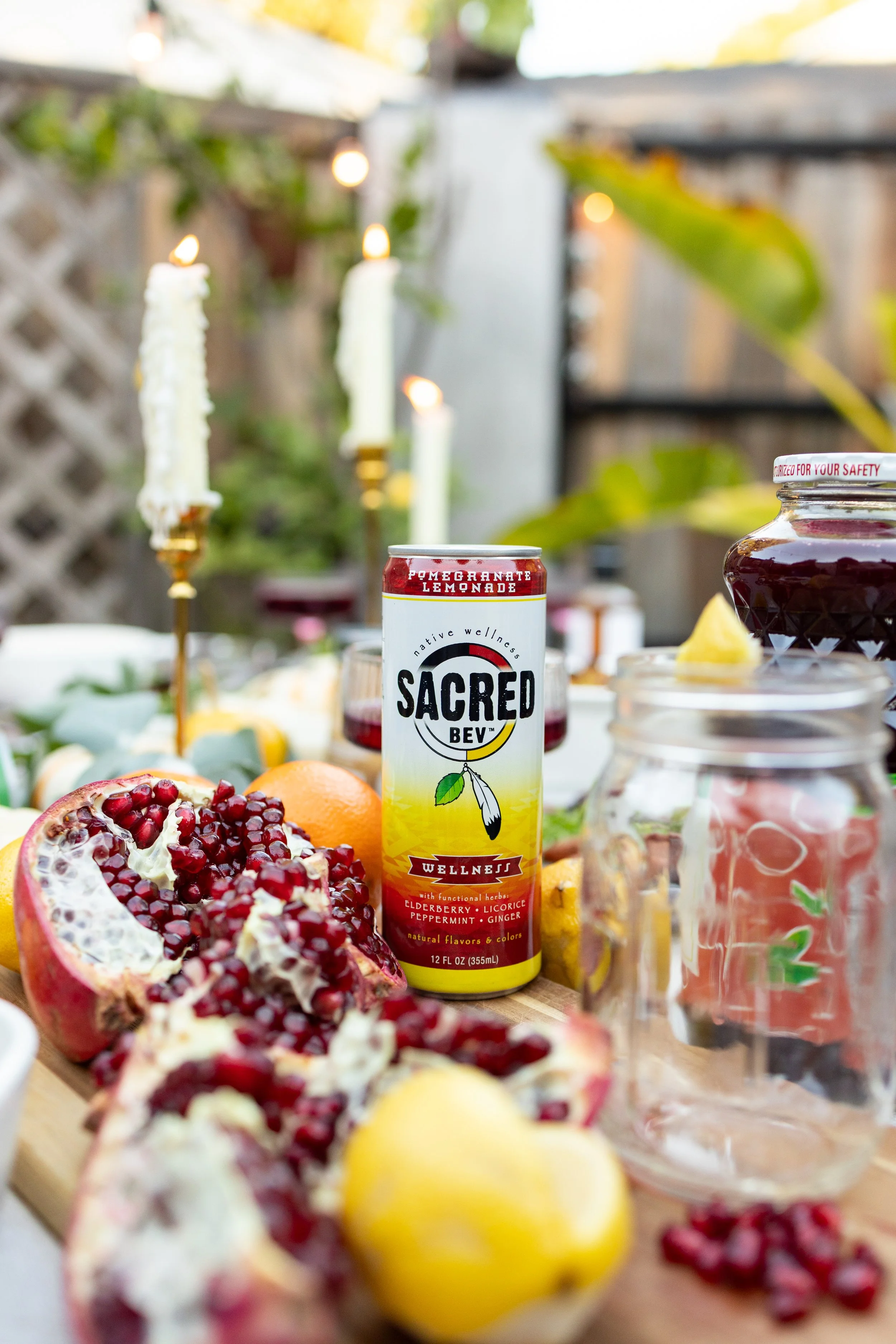 A drinks table featuring a can of Sacred Wellness pomegranate lemonade, pomegranate, lemons, glass jars with drinks, a jar with a lemon wedge, and candles in an outdoor setting.