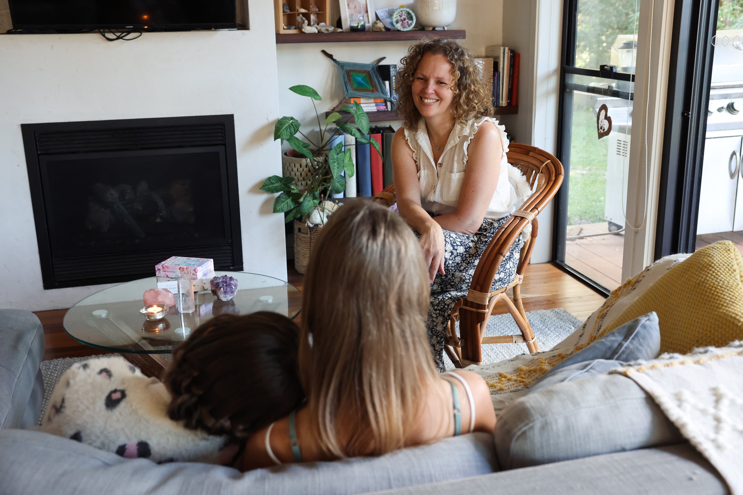 A woman with curly hair sitting in a chair smiling at two children lying on a couch, with a fireplace, a glass coffee table with candles and crystals, and a sliding glass door in the background.