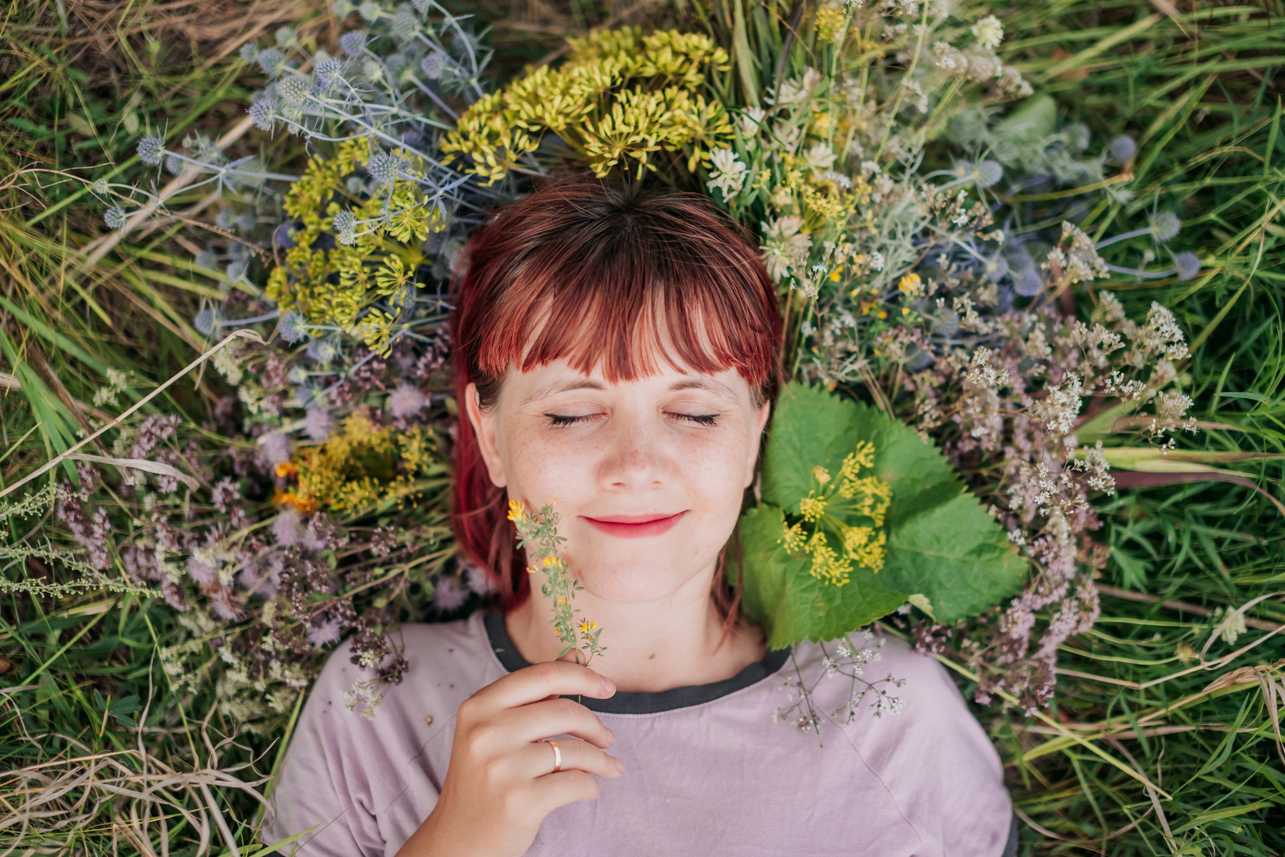 A woman with red hair and closed eyes lying on ground surrounded by green grass and various wildflowers, holding a small yellow flower near her face.