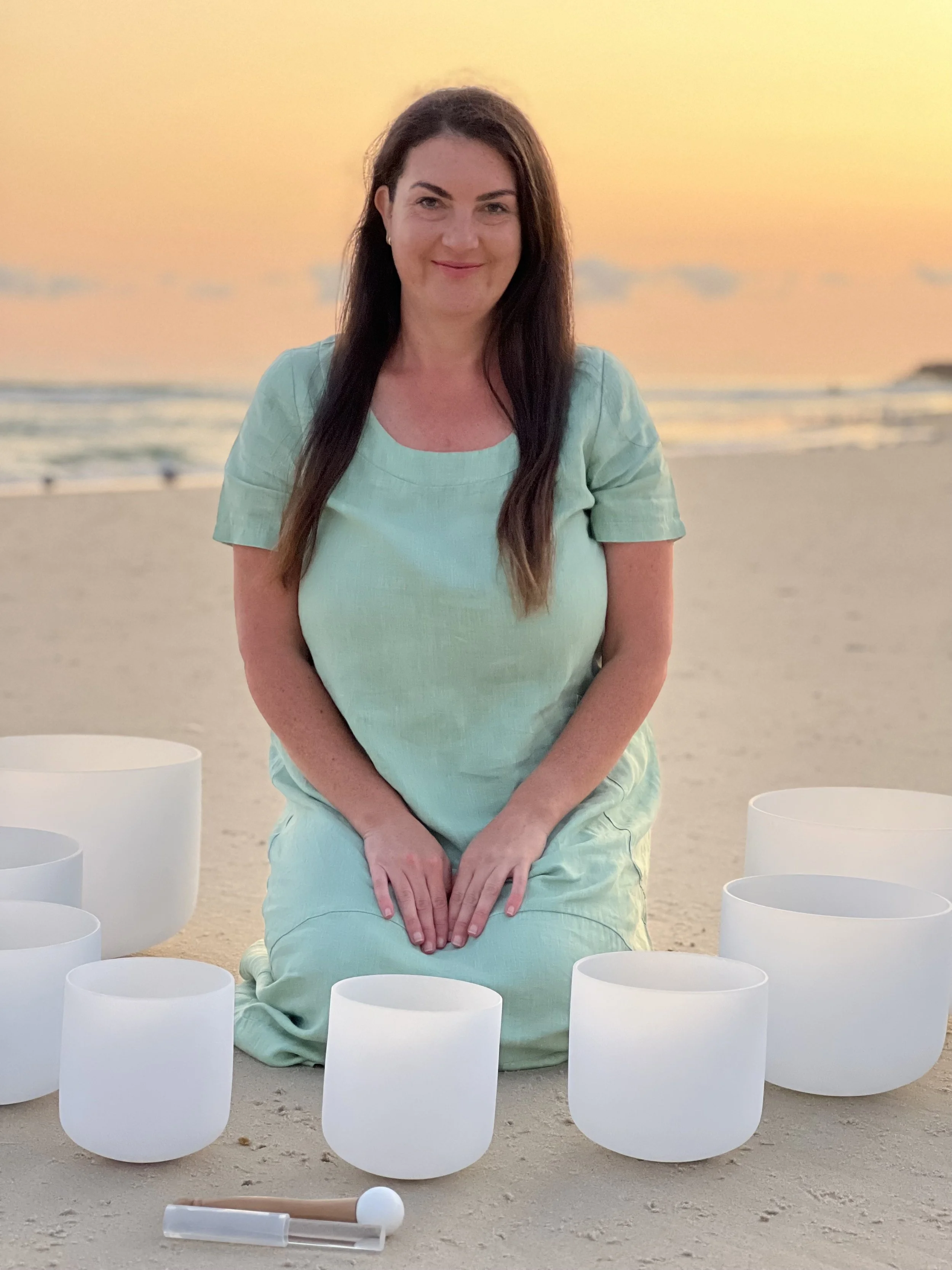 Woman sitting on a sandy beach at sunset with white crystal singing bowls around her.