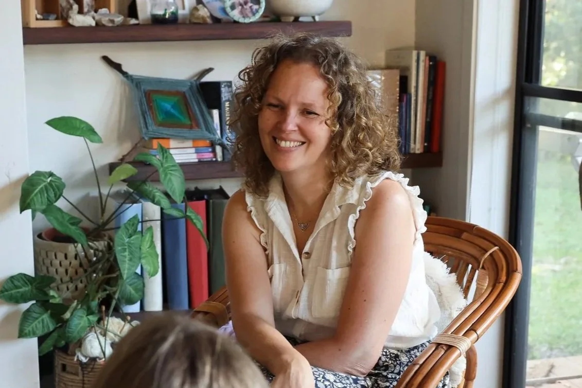 A woman with curly hair smiling and sitting on a wicker chair in a room with books, a plant, and a window.