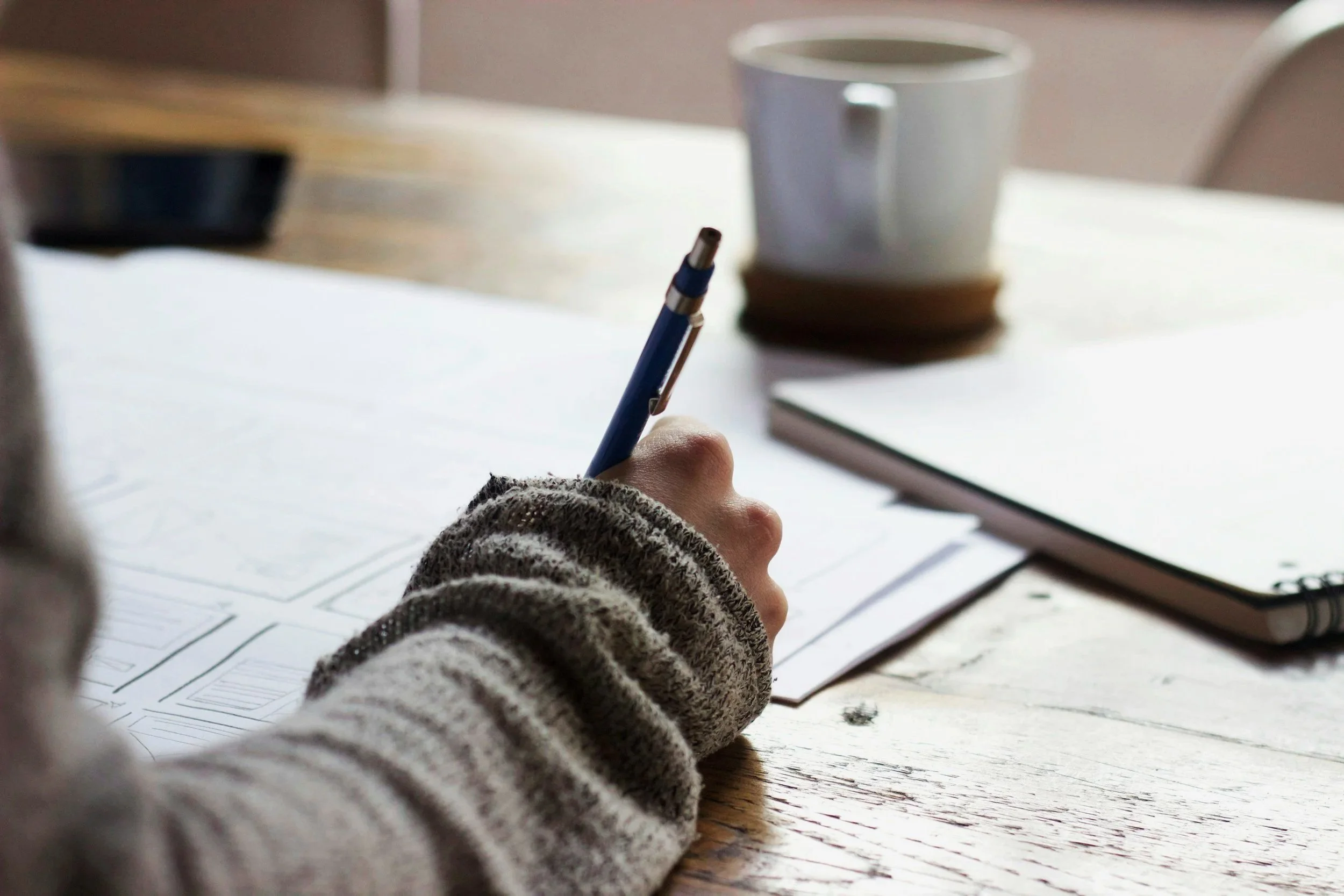 Person writing on papers with a blue pen on a wooden table, with a notebook and a white mug in the background.