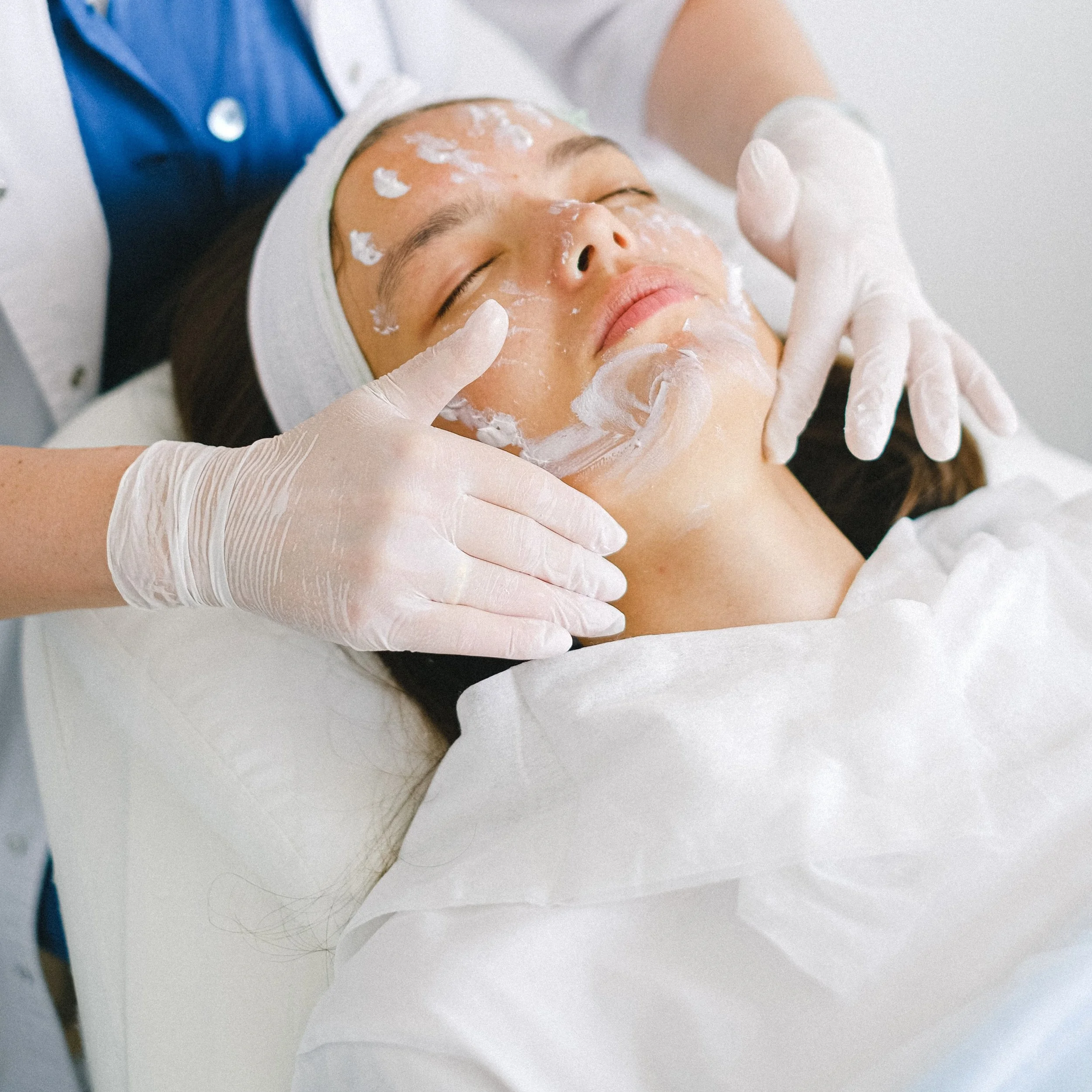 A woman lying down with her eyes closed, receiving a skincare treatment, with a facial mask applied, in a clinical setting.