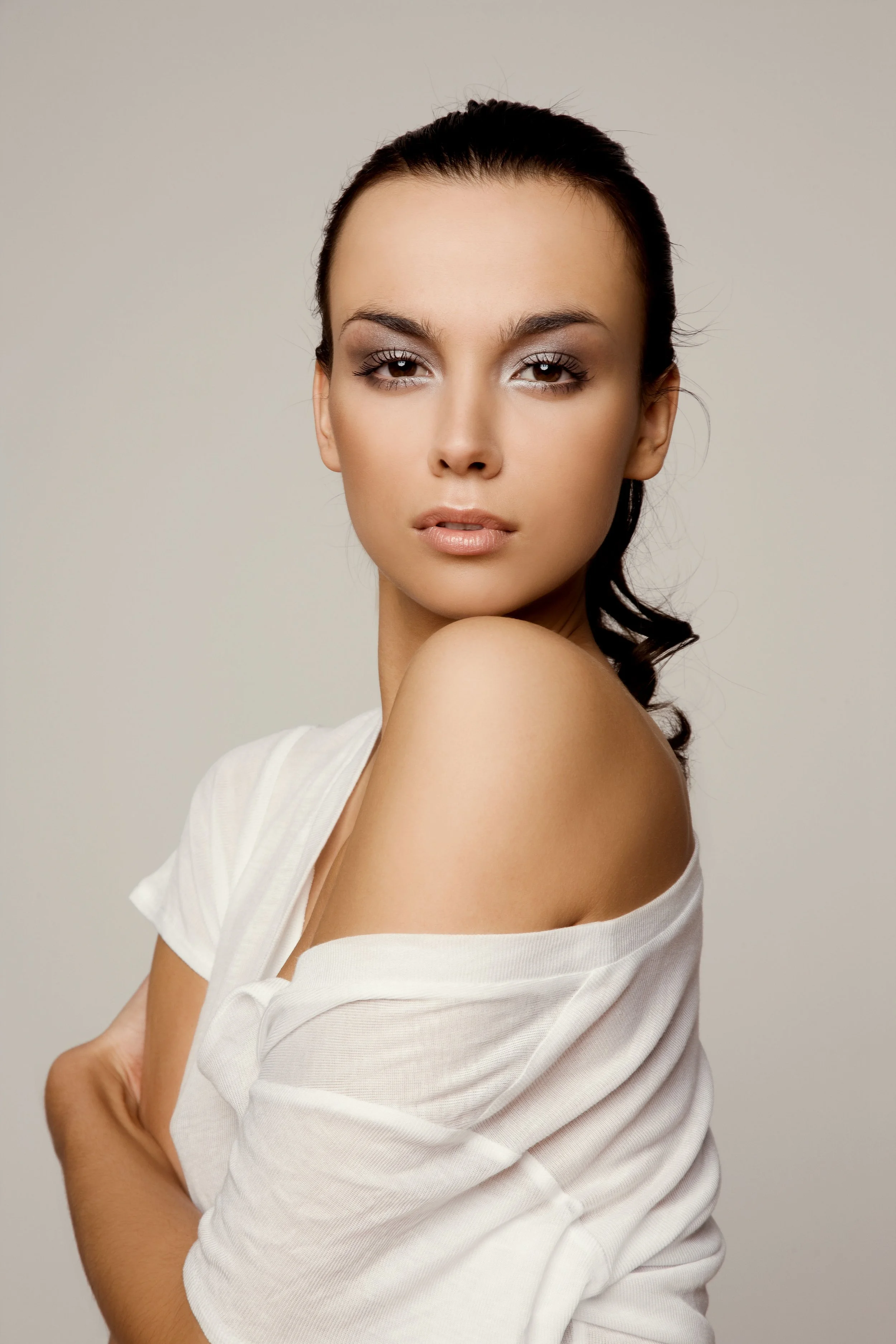A woman with dark hair pulled back, wearing a white off-the-shoulder top, looking at the camera with a neutral expression
