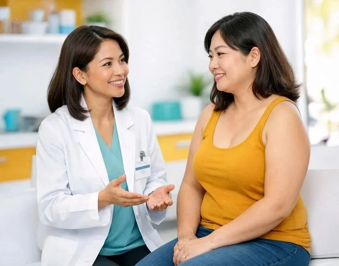 A female doctor in a white coat talking to a patient in a yellow tank top during a consultation in a medical office.