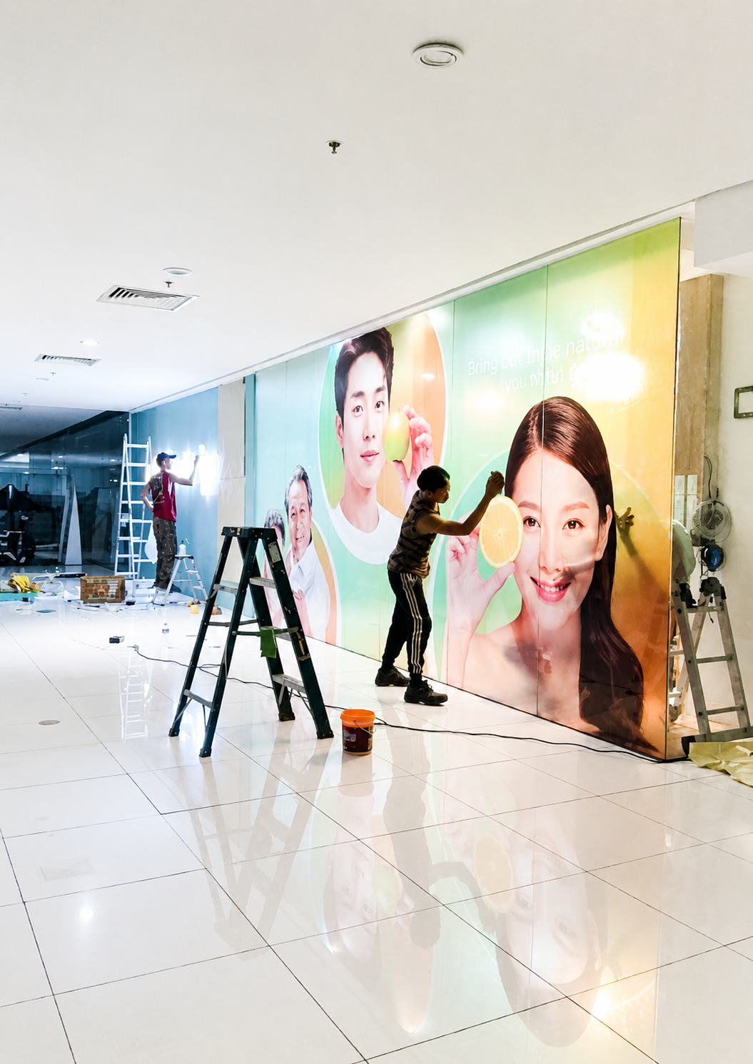 Workers installing a large colorful advertisement featuring people holding citrus fruits inside a building.