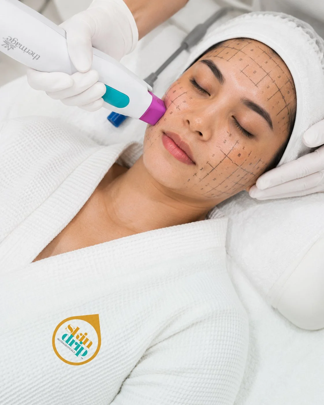 A woman receiving a facial or skin treatment with a handheld device on her face. She is lying down with her eyes closed, wearing a white headband and a white robe. The face has markings for cosmetic procedures and there is a logo on her robe that says 'skin drip'.