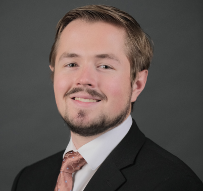 A professional man in a black suit and pink tie smiling against a dark gray background.