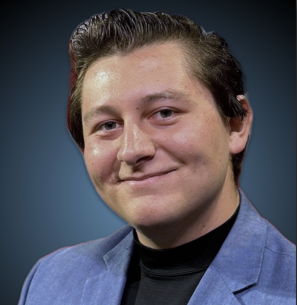 A young man with dark hair and fair skin smiling in front of a U.S. flag, wearing a blue blazer, a black turtleneck, and a name badge reading "Lake Dodson".