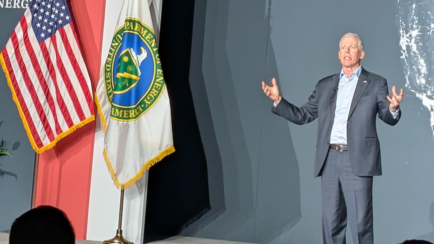 Chris Wright speaking on stage with US, DOE flags in the background.