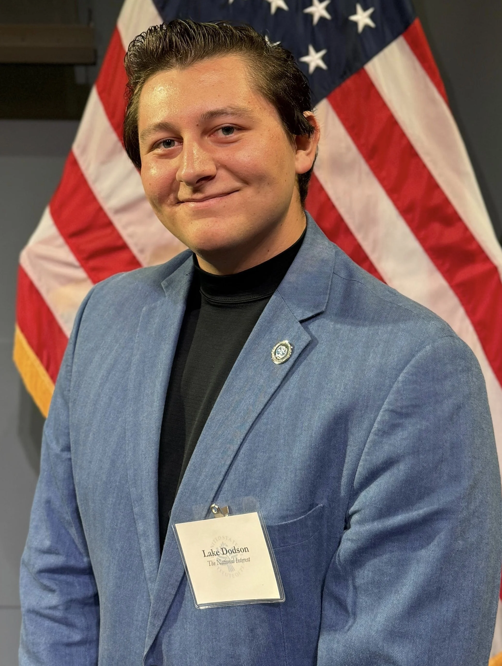 Young man in a blue blazer, black turtleneck, and a name badge standing in front of an American flag.