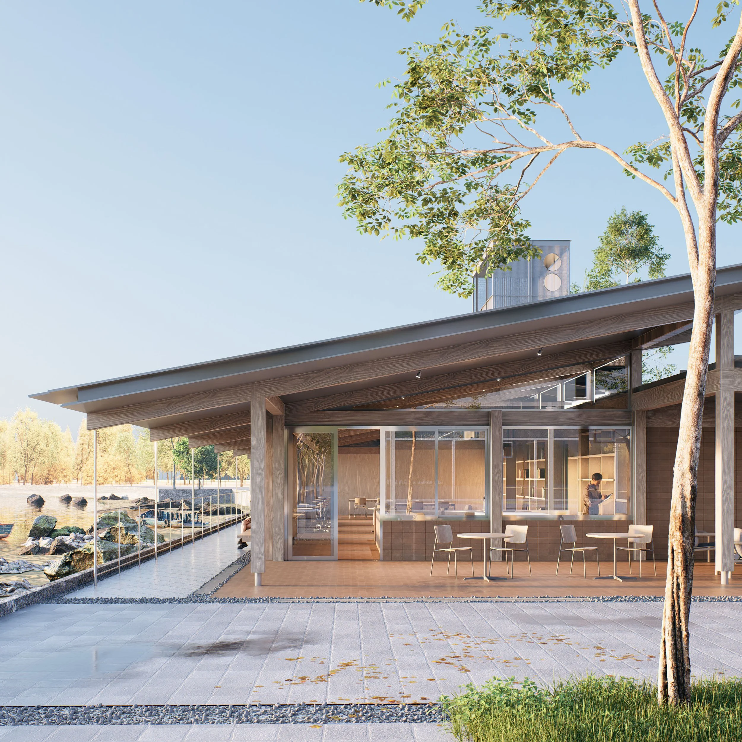 Modern glass-fronted building with outdoor seating near a rocky shoreline, trees, and a clear blue sky.