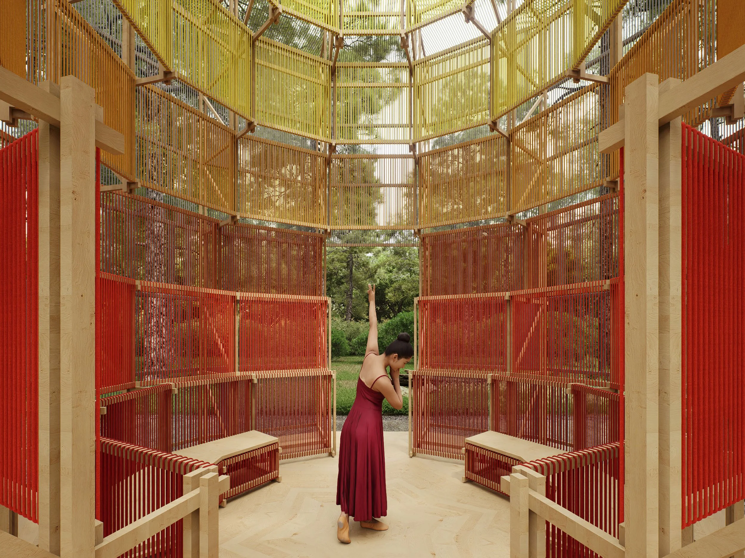 A woman in a maroon dress performing ballet inside a colorful, open-air wooden structure with red, yellow, and orange vertical slats, set in a park with green trees in the background.