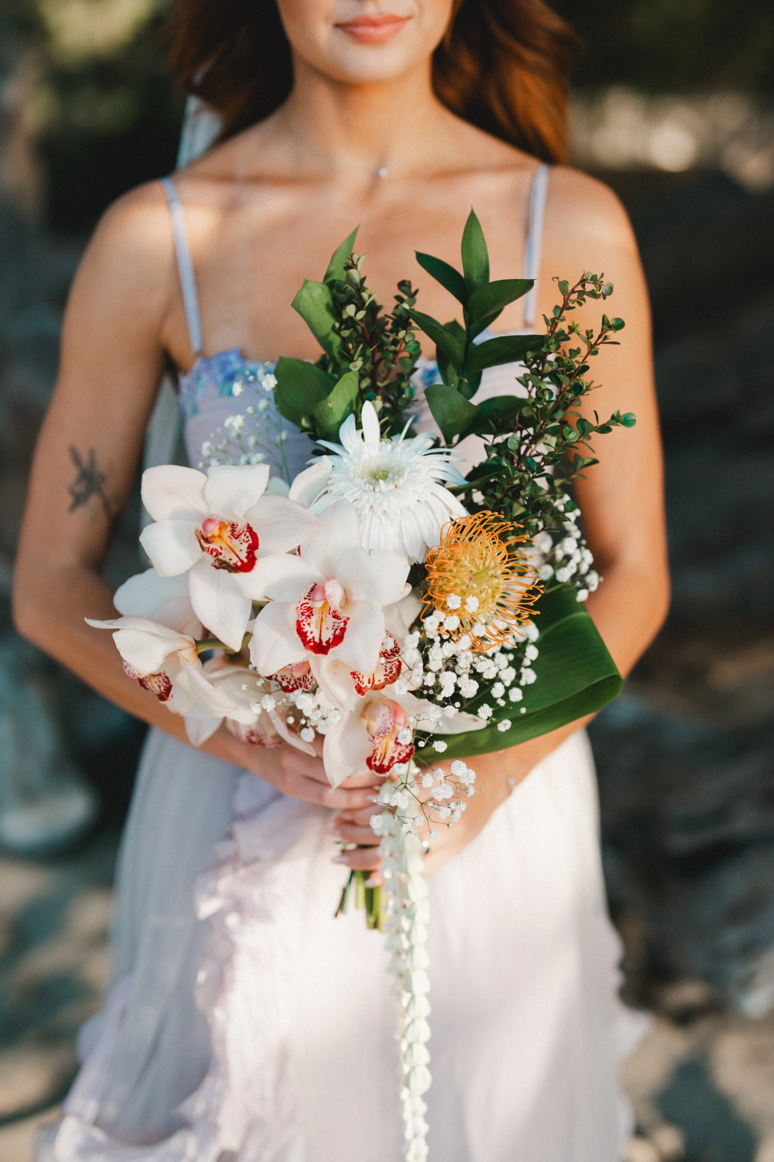 A woman in a white dress holds a bouquet of tropical flowers, including orchids, greenery, and other vibrant blossoms, outdoors.