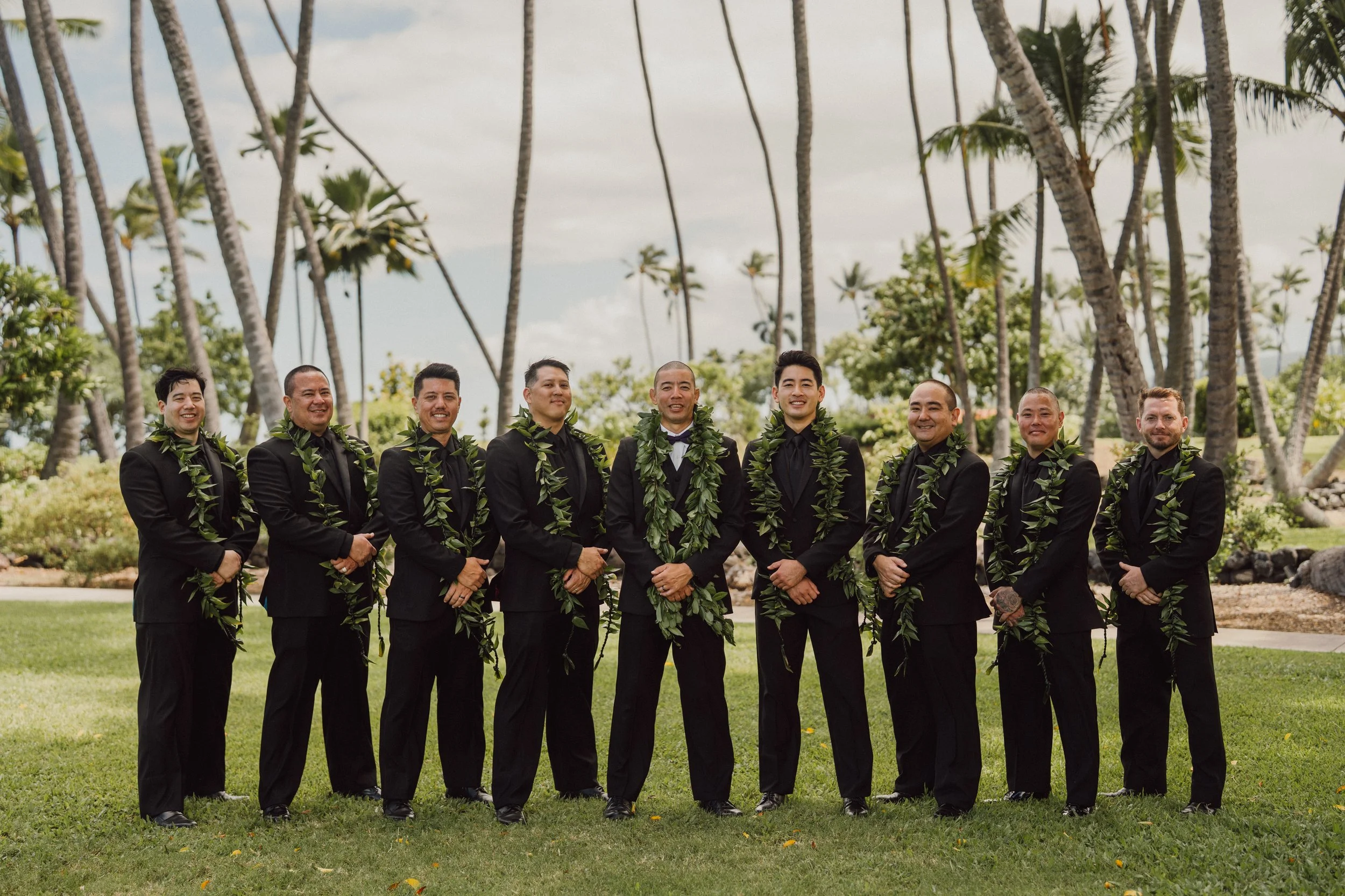 A group of nine men dressed in black suits with leis around their necks, standing outdoors on grass with palm trees in the background.
