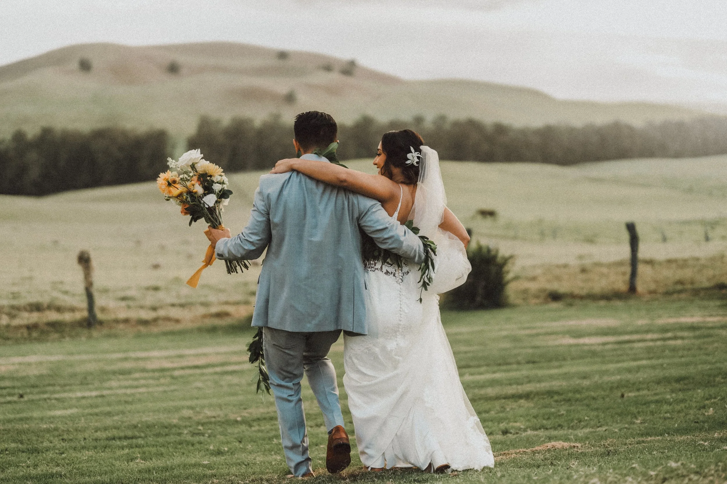 A bride and groom walking arm in arm on a grassy field, with rolling hills in the background, during their wedding celebration.