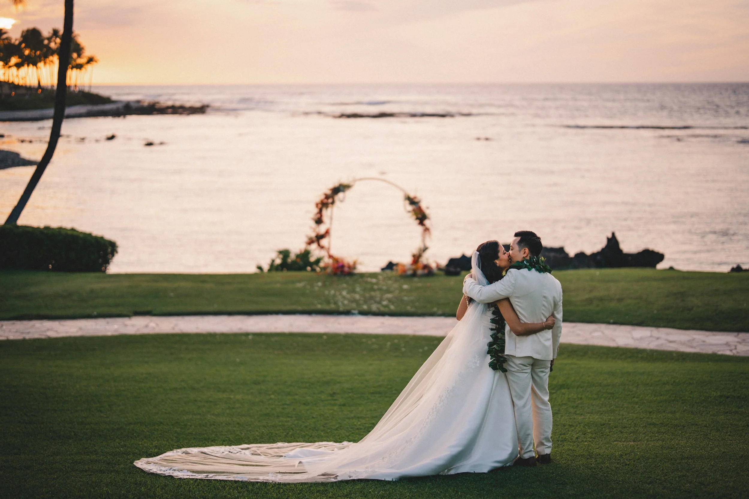 A bride and groom sharing a kiss on a lawn by the ocean during sunset, with an arched floral wedding decoration in the background.