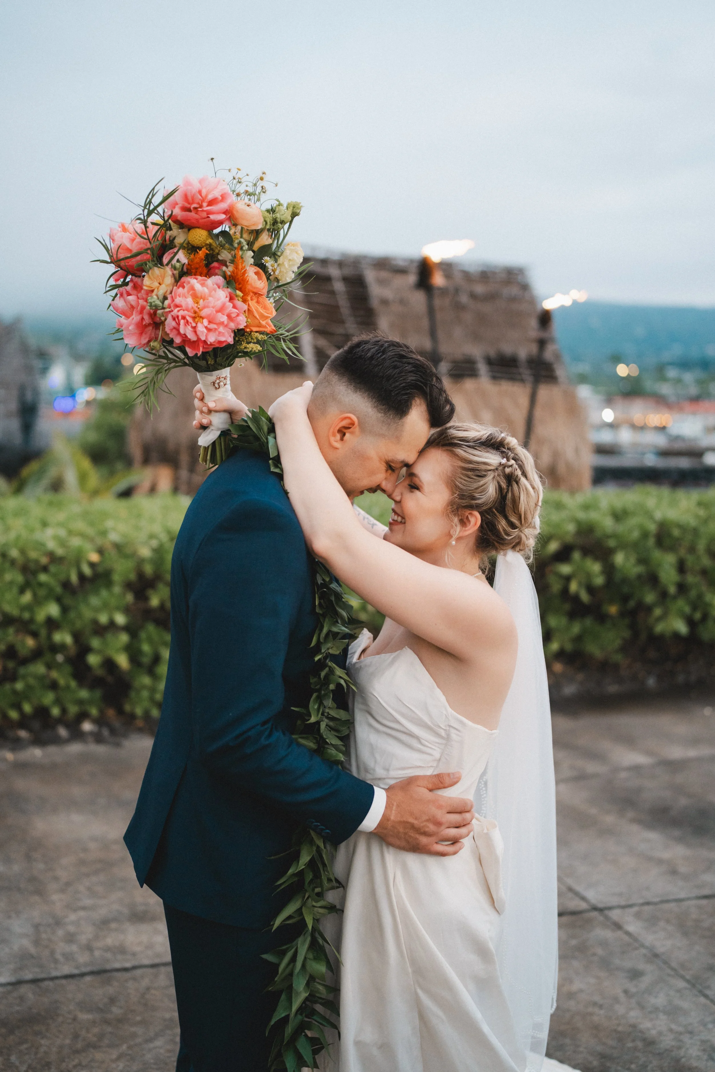 A newlywed couple embraces, smiling and touching foreheads, outdoors with a thatched-roof building in the background at dusk. The groom wears a navy suit and a greenery lei, holding a bouquet of pink and orange flowers. The bride is in a white strapl