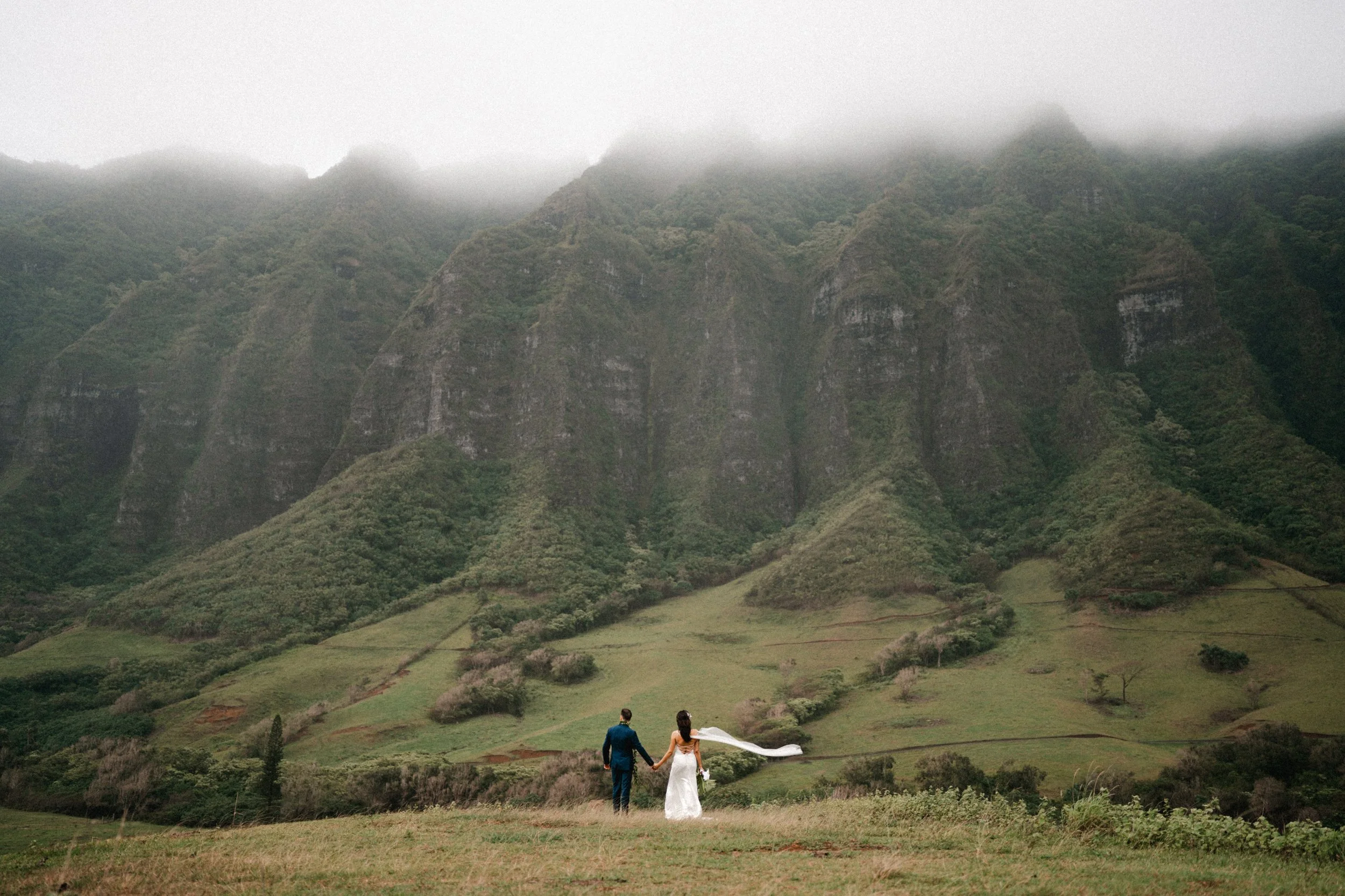 A couple in wedding attire walking hand in hand in a grassy field with lush green hills and mountains in the background, partially covered by fog