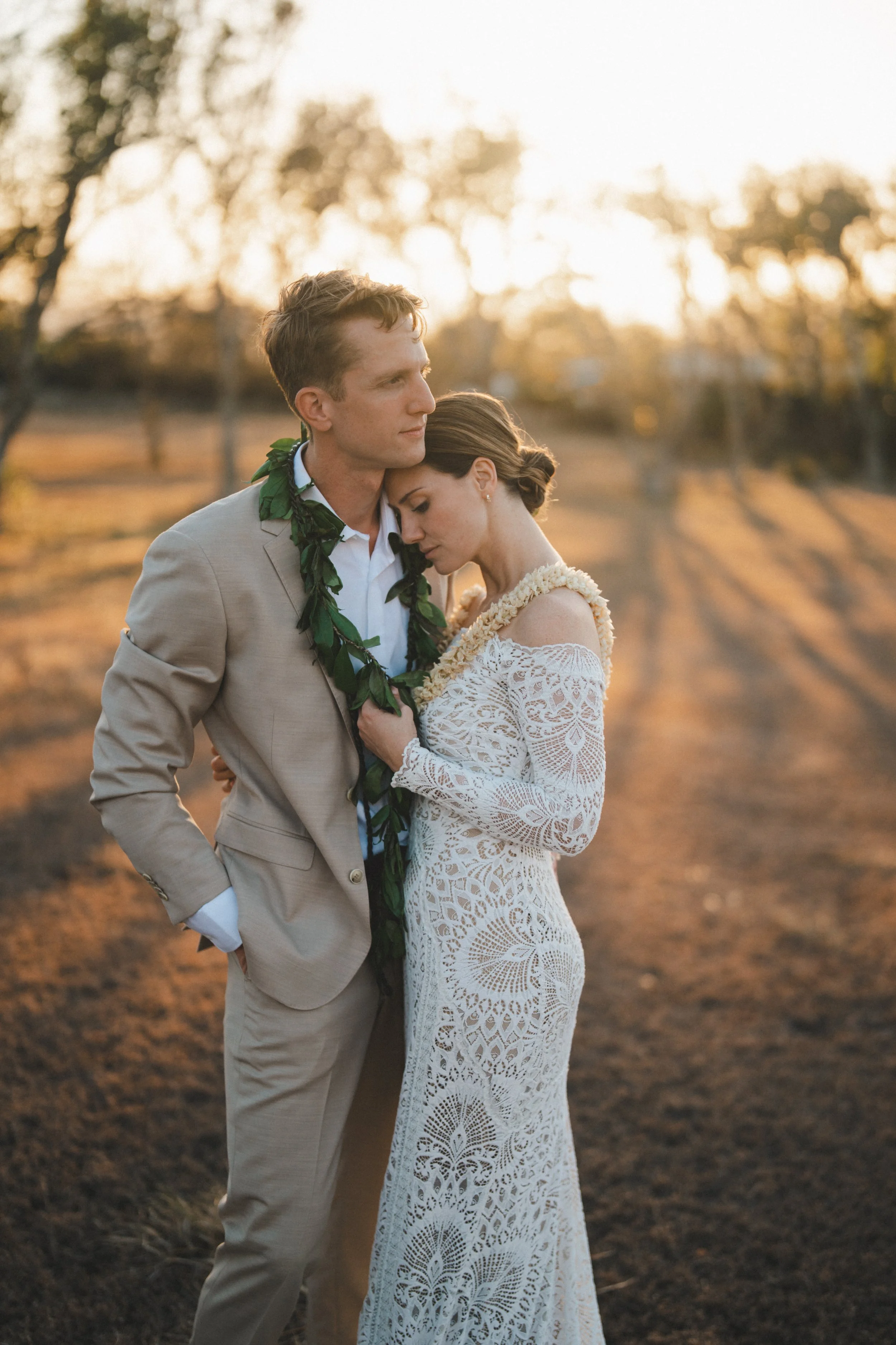 A couple in wedding attire embraces in a field at sunset, with trees in the background.