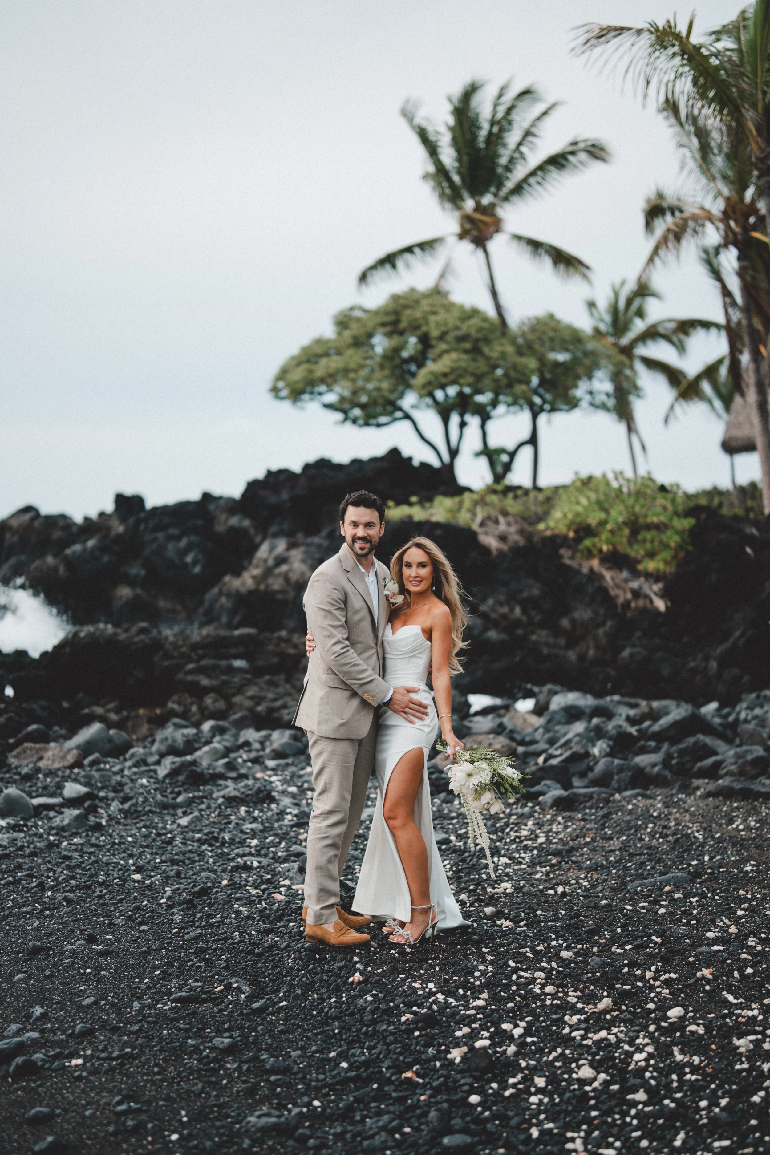 A couple in wedding attire standing on a black sand beach with palm trees and volcanic rocks in the background.