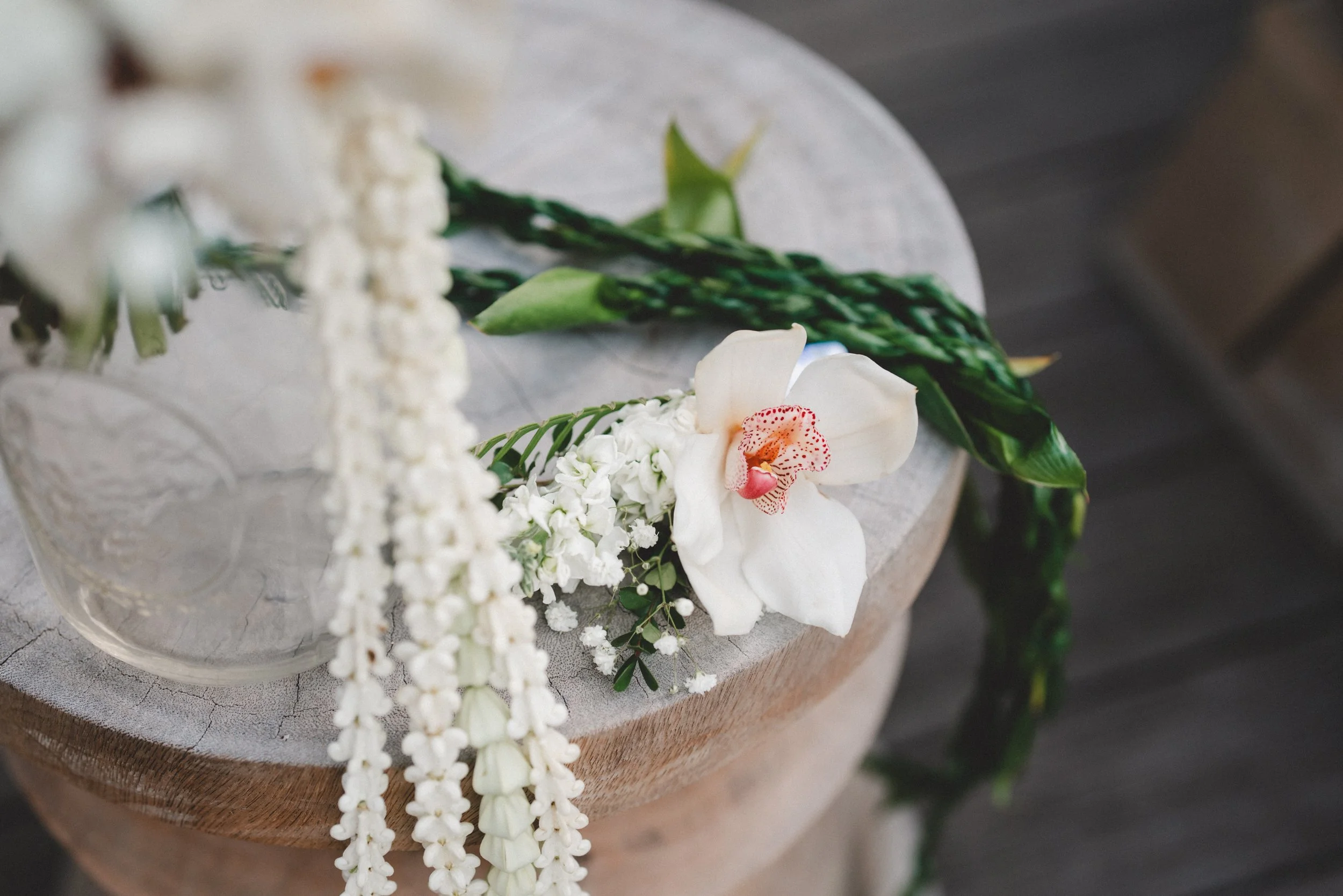 A floral arrangement with a white orchid, white cascading flowers, and green foliage on a round wooden table.