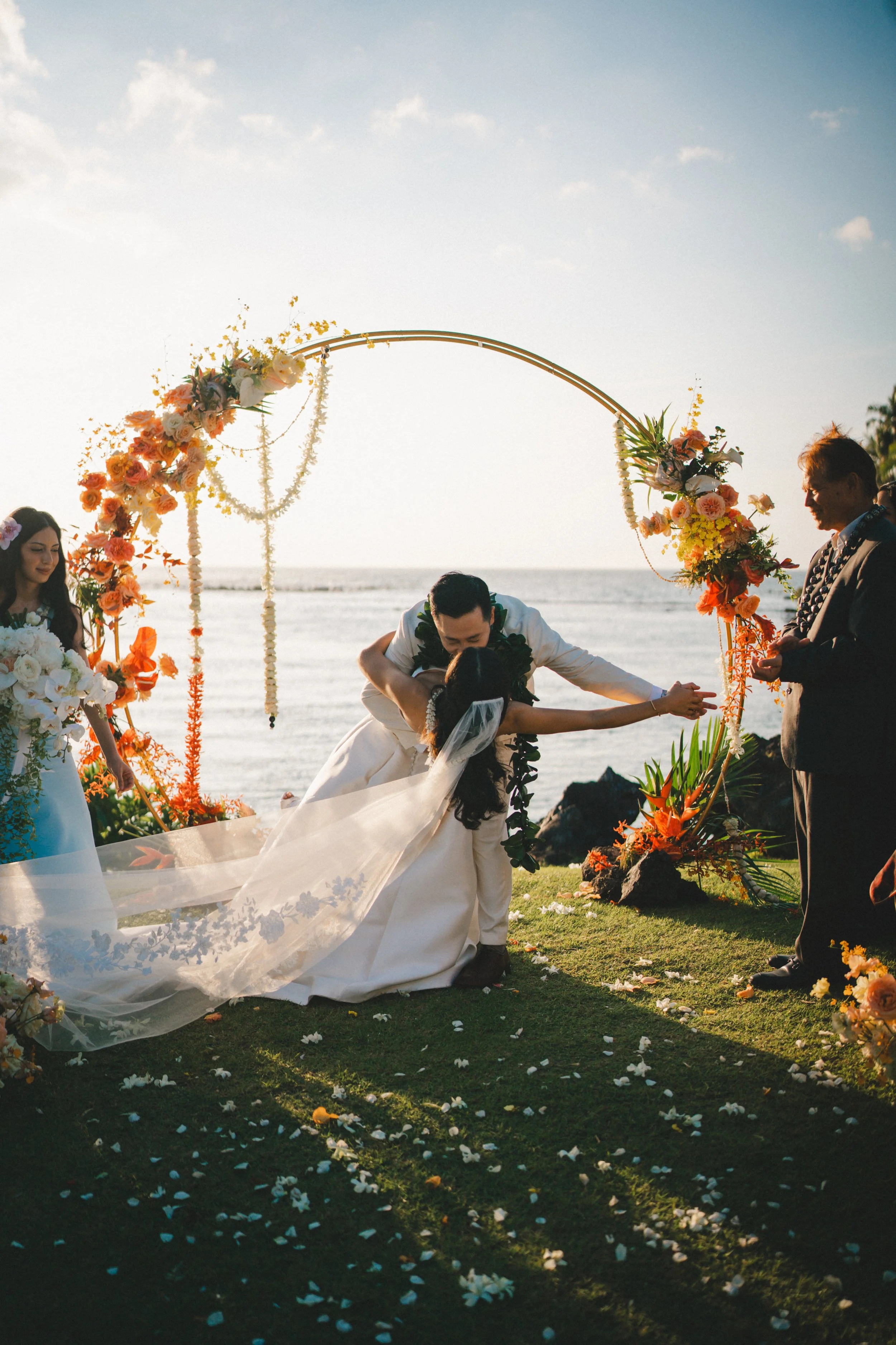 A bride and groom kissing under a floral arch during an outdoor beach wedding at sunset, with a bridesmaid and officiant nearby.