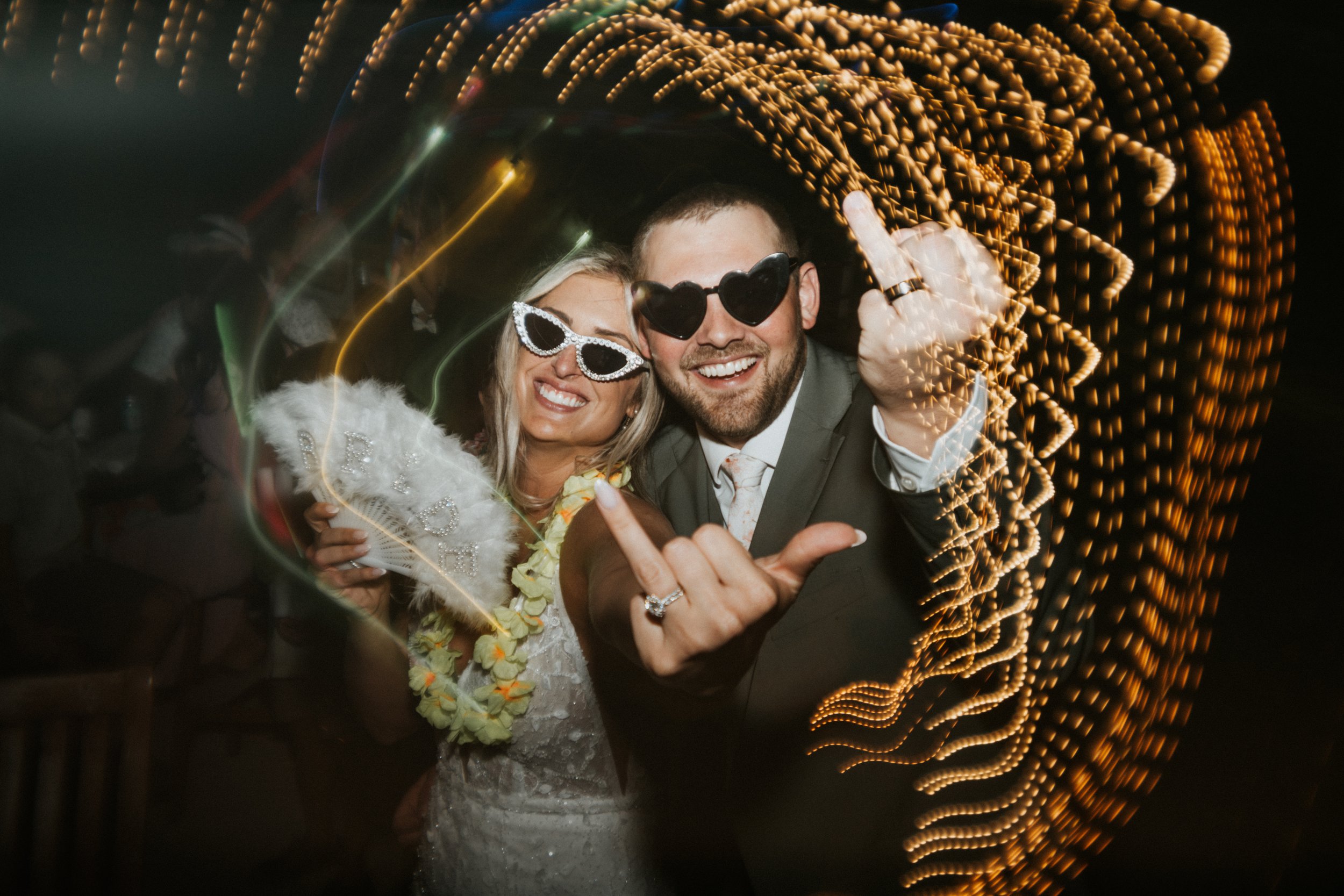 A lively photo of a smiling couple at a celebration, wearing sunglasses, with colorful lights creating a swirl effect around them.