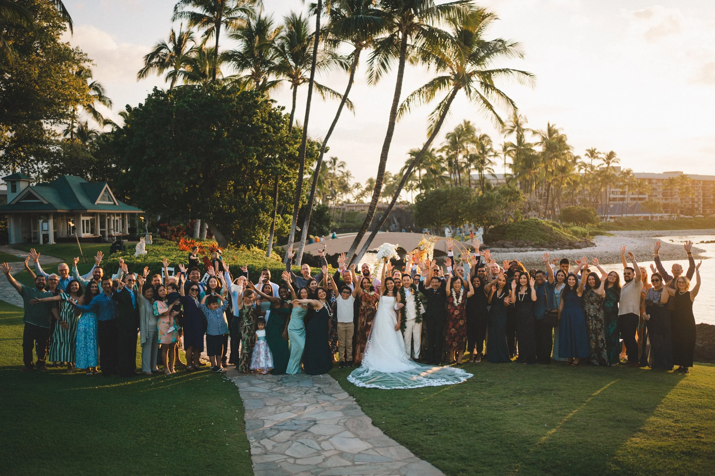 A large group of people celebrating a wedding outdoors near a beach with palm trees during sunset.