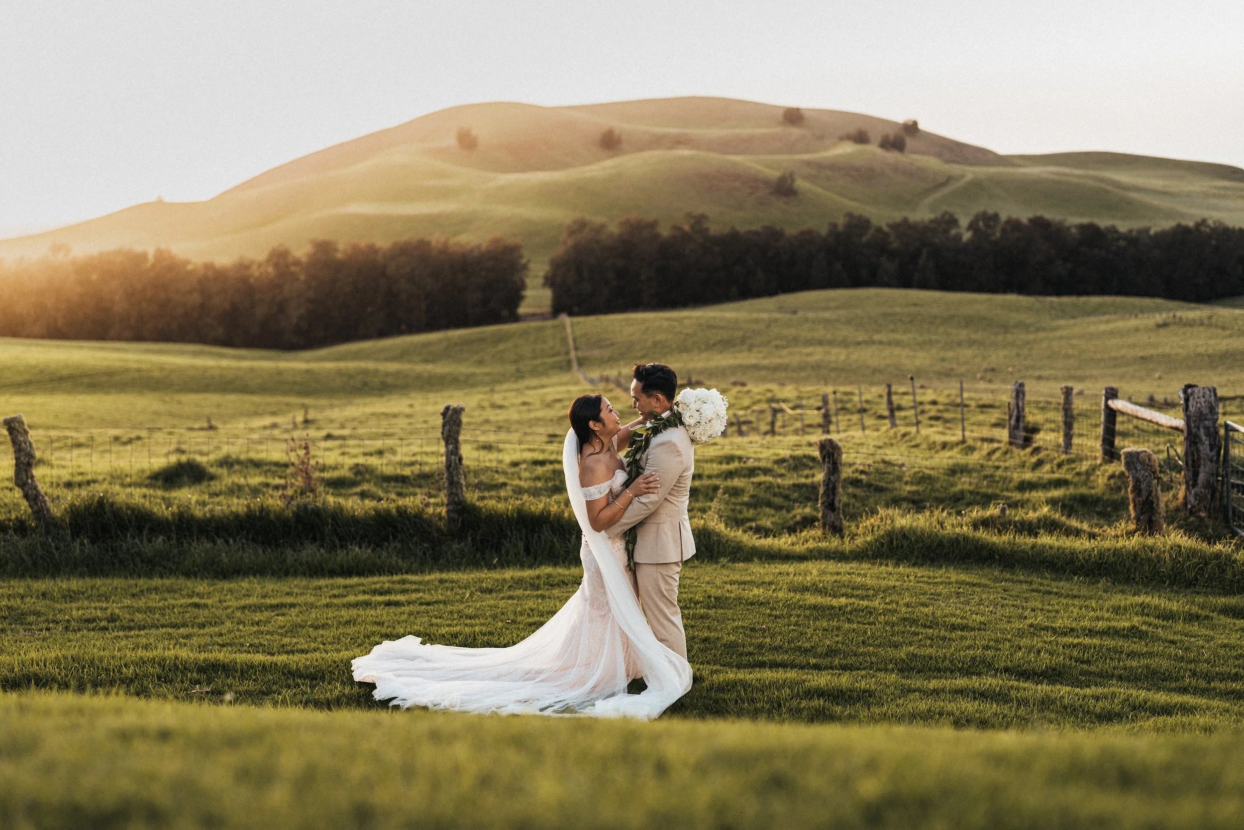 A bride and groom in wedding attire standing close and smiling in a lush green field during sunset, with rolling hills and a fence in the background.