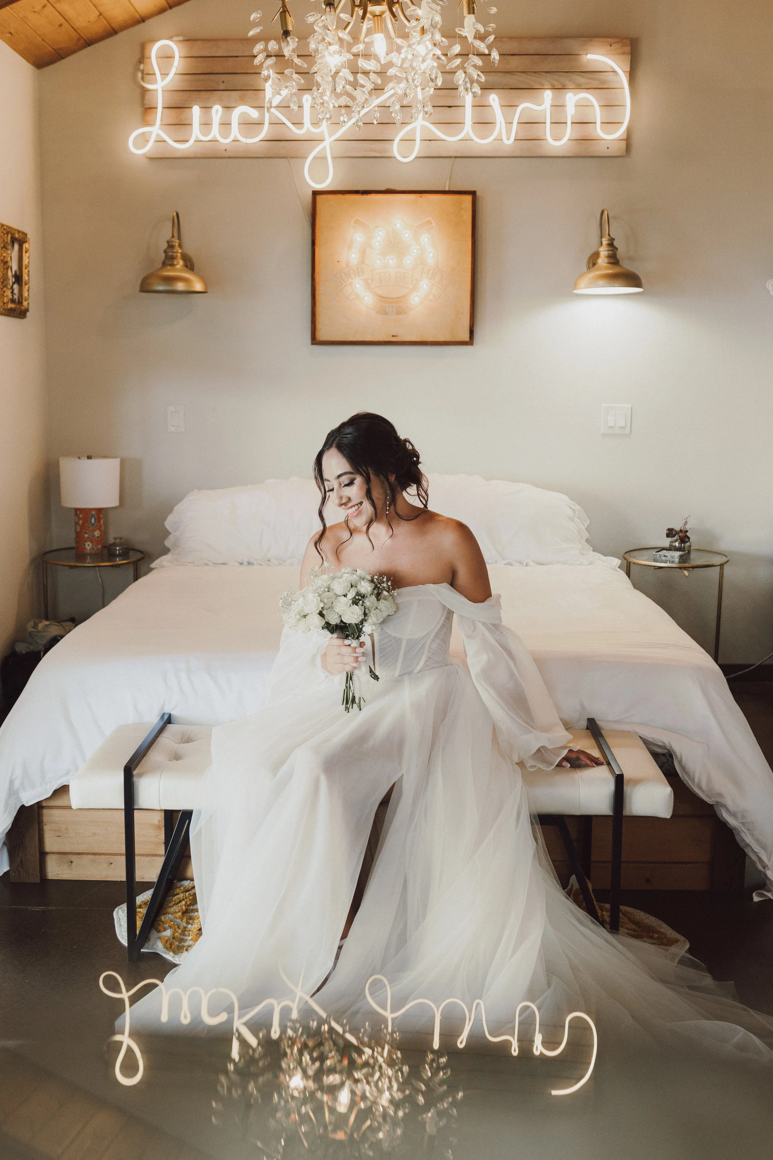 A bride in a white wedding dress holding a bouquet of flowers sitting on a bench in a bedroom, with a neon sign that says 'Lucky Girl' on the wall behind her.